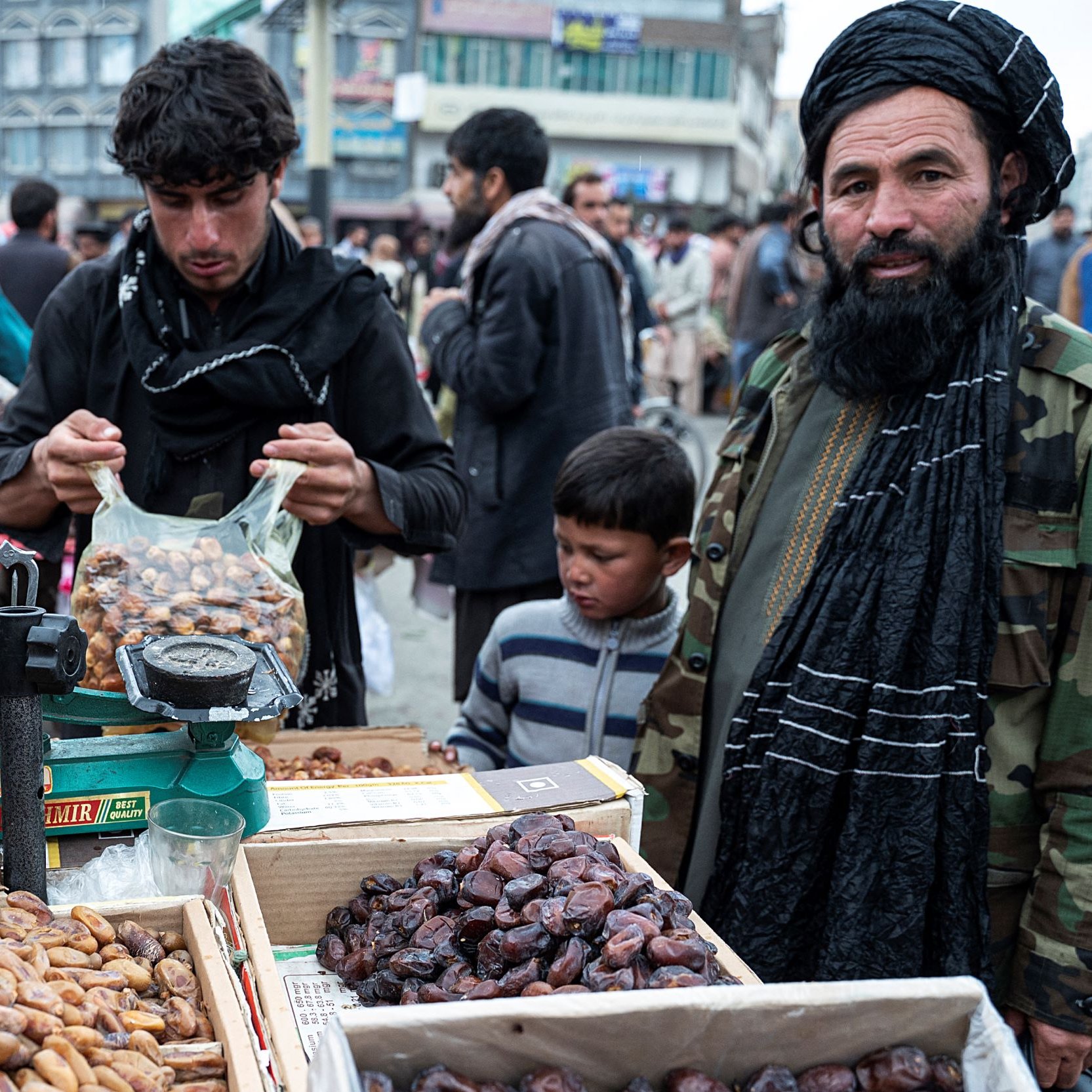 The image depicts a bustling market scene, likely in Afghanistan. In the foreground, there is a vendor displaying various types of dates in baskets and boxes. The vendor, dressed in a traditional outfit and a black turban, stands next to a scale. Another young man is shown packing dates into a bag, while a young boy looks on. The background features a crowd of people, creating a lively atmosphere, with buildings visible in the distance. The overall scene reflects daily life and commerce in a market setting.