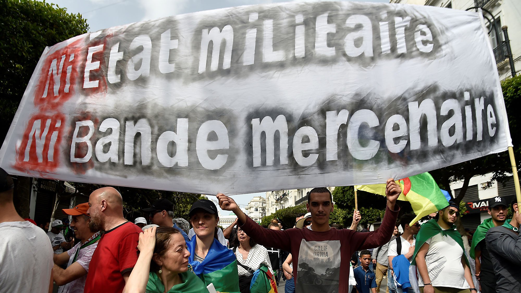 L'image montre une manifestation où des gens tiennent une grande banderole. Sur celle-ci, on peut lire "Ni État militaire, ni Bande mercenaire", ce qui reflète un message de revendication politique. Les manifestants semblent porter des drapeaux et exprimer des sentiments de dissentement. L'ambiance est mobilisée, avec des personnes de différents âges rassemblées pour une cause commune.
