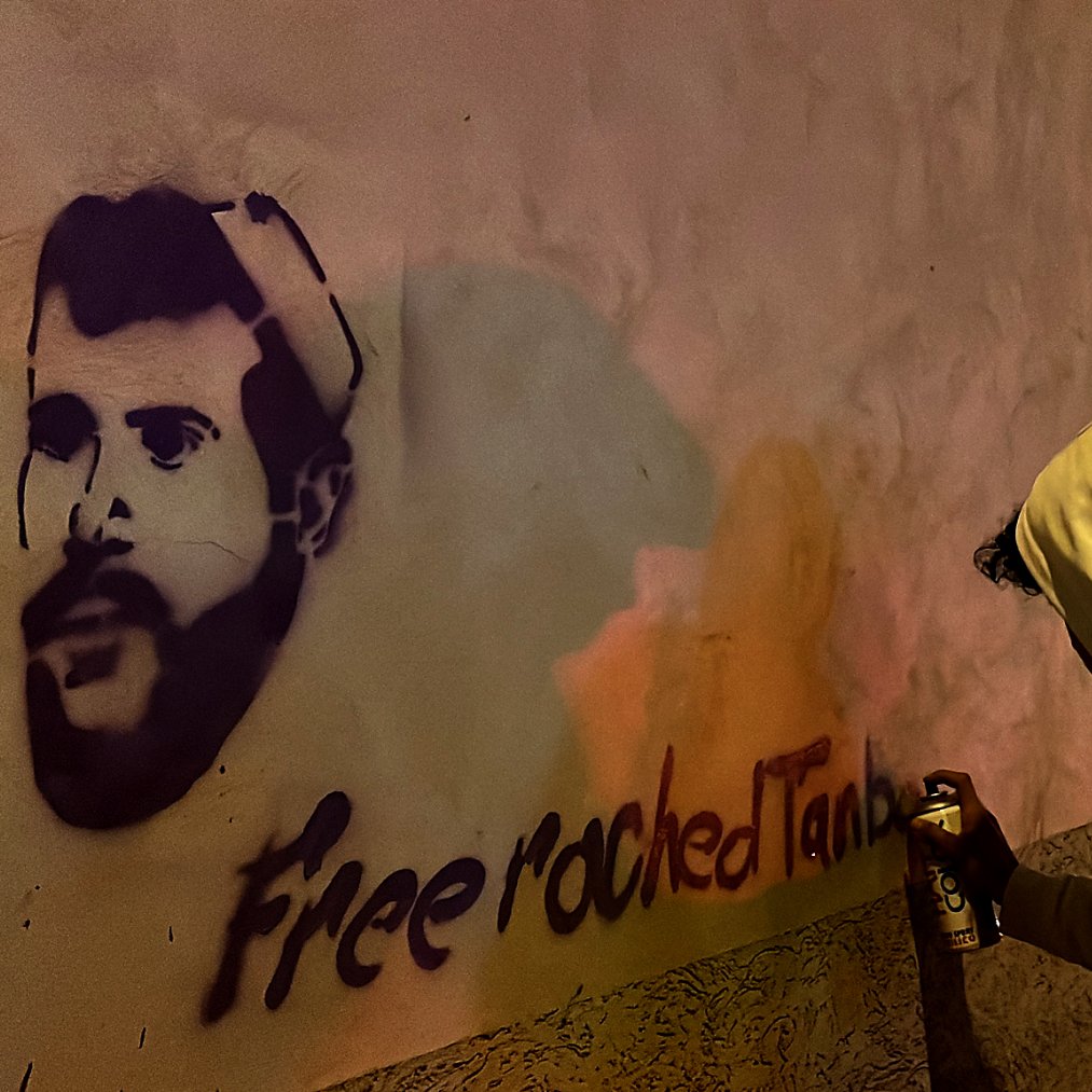 تظهر في الصورة شخص يرتدي سترة هوديي بيضاء وهو يقوم برسم جرافيتي على حائط. يظهر على الحائط رسم لصورة شخصية، إلى جانب عبارة "Free roched Tani" مكتوبة بشكل جرافيتي. تبدو الخلفية مظلمة، مما يعكس أجواء الاحتجاج أو النشاط الفني في الليل.