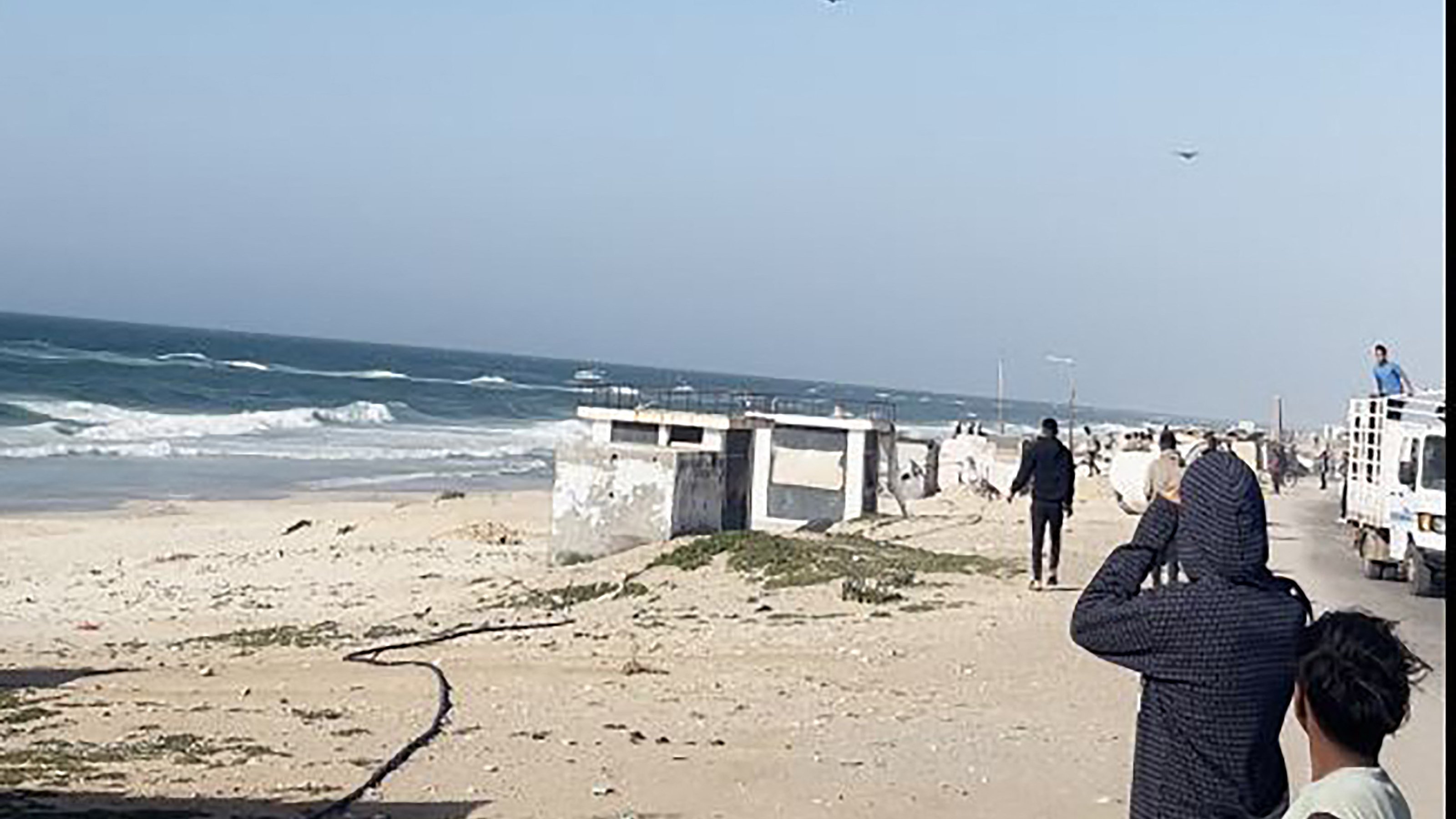 L'immagine mostra una scena costiera con il mare e delle onde in movimento. Si possono vedere alcune persone sulla spiaggia che osservano il mare, apparentemente interessate a qualcosa in cielo. Il paesaggio è caratterizzato da sabbia chiara e una scalinata che porta a una struttura, mentre in lontananza si vedono veicoli e persone che si muovono lungo la costa. Il cielo è sereno, con poche nuvole.
