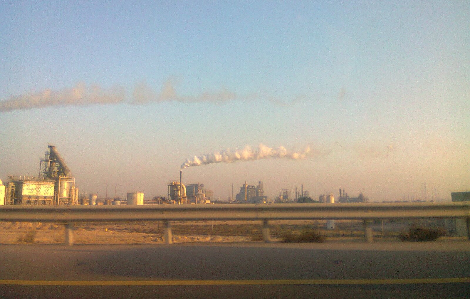 The image depicts an industrial landscape with several factories. In the foreground, there is a highway barrier, suggesting the viewer is looking from a moving vehicle. The factories emit smoke and steam into the atmosphere, creating white clouds against a clear blue sky. The overall scene conveys an industrial environment, typical of urban or semi-urban areas where manufacturing activities take place.