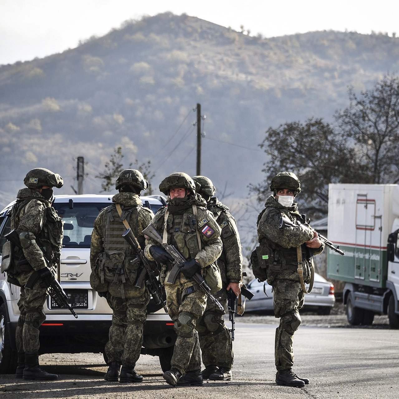 تصویر گروهی از سربازان مسلح را نشان می‌دهد که در یک منطقه روستایی قرار دارند. سربازان در لباس‌های نظامی و کلاه‌های ایمنی به وضوح مسلح هستند و در حال برقراری امنیت به نظر می‌رسند. پس‌زمینه تصویر شامل کوه‌ها و درختان است که به فضا حالت طبیعی می‌دهد. همچنین، چندین وسیله نقلیه در کنار جاده دیده می‌شوند. نور تصویر نیز نشان‌دهنده روز روشن است.