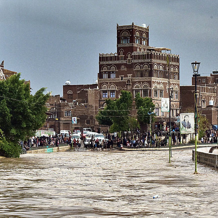 L'image montre une scène de inondation dans une ville, probablement au Yémen. On peut voir des bâtiments traditionnels en pierre avec des arches et des fenêtres ornementales. Les niveaux d'eau sont élevés, couvrant une grande partie de la rue, ce qui semble indiquer des pluies abondantes. Des gens se rassemblent sur le bord, observant la situation. Le ciel est nuageux, ajoutant à l'atmosphère dramatique de la scène. Les arbres sont verdoyants, indiquant une nature environnante peu affectée par la montée des eaux.
