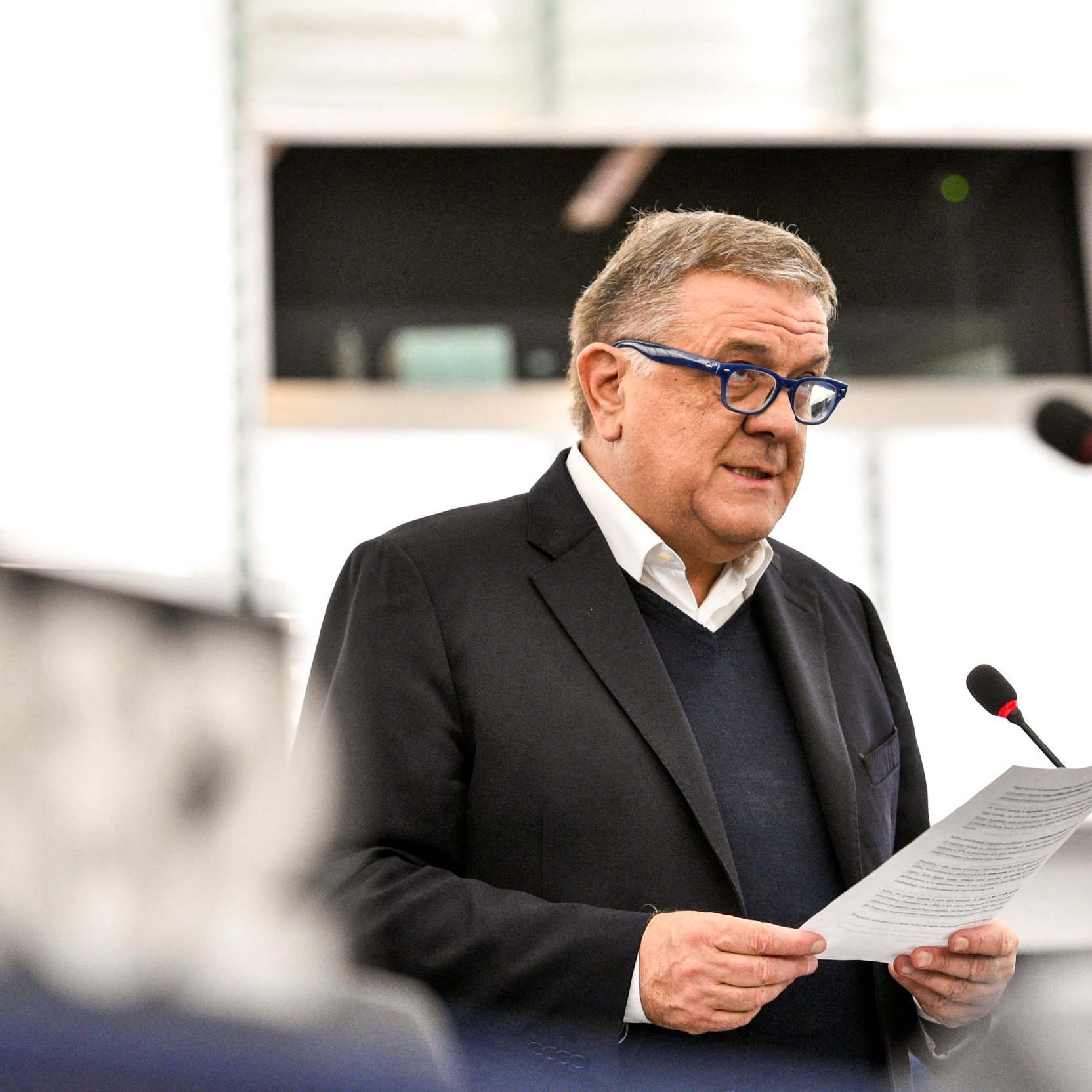 L'image montre un homme qui parle au micro lors d'une session au Parlement. Il porte des lunettes bleues et un costume sombre. Il semble lire un discours ou un document. L'environnement est celui d'une salle de réunion institutionnelle, avec un éclairage moderne et des panneaux en arrière-plan.