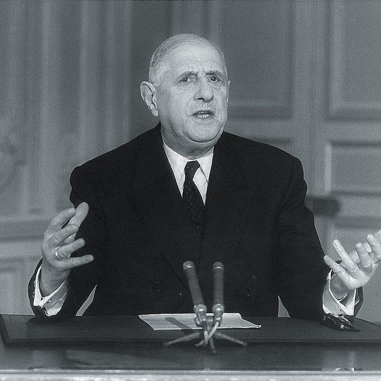 L'image montre un homme en costume formel, assis à une table avec un microphone devant lui. Il semble être en train de parler ou de faire un discours, utilisant des gestes expressifs avec ses mains. L'arrière-plan est sobre, avec des murs ornés de motifs simples. L'éclairage met en valeur son visage, soulignant son sérieux et son engagement dans son discours.