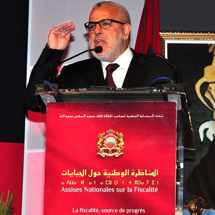 L'image montre un homme en train de s'exprimer lors d'un événement officiel. Il porte des lunettes et une veste noire, et il semble faire un geste de salutation avec sa main. Derrière lui, un panneau avec du texte en arabe et français est visible, ainsi qu'une photo d'un autre homme, probablement une figure politique. Le contexte semble être lié à des discussions sur des questions fiscales.