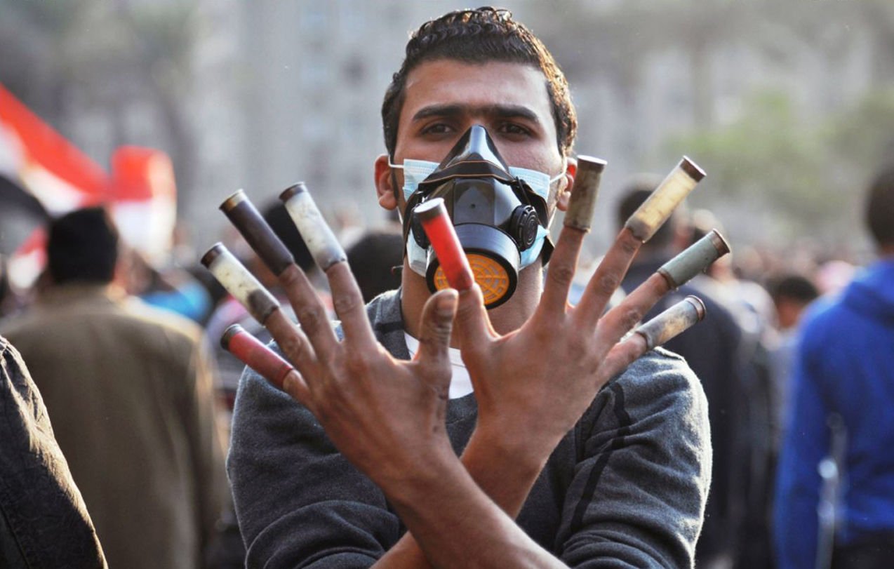 L'image montre un homme portant un masque de protection, levant les mains avec plusieurs cartouches de gaz lacrymogène visibles. Son expression semble déterminée, et l'arrière-plan indique qu'il est présent dans une foule, probablement lors d'une manifestation. L'atmosphère de la scène semble être celle d'un événement intense et chargé d'émotion.