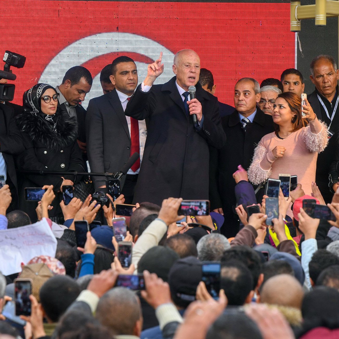 L'image montre un groupe de personnes rassemblées devant une scène où un homme, probablement un leader politique, prend la parole. Il est entouré de plusieurs personnes, dont des membres de la sécurité et des journalistes avec des caméras. La foule est très engagée, beaucoup de gens tiennent des téléphones pour filmer ou prendre des photos. L'arrière-plan présente des affiches ou des bannières, probablement en rapport avec un événement important. L'ambiance semble dynamique et pleine d'énergie.