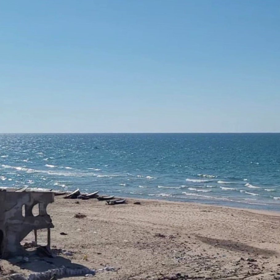 L'image montre une plage sous un ciel bleu clair, avec une mer d'un bleu éclatant qui s'étend à perte de vue. Le rivage est bordé de sable fin, mais des débris, comme une structure en béton en ruine, sont visibles sur la plage, ajoutant un contraste à la beauté naturelle du paysage. Au loin, on peut apercevoir quelques petites embarcations sur l'eau, suggérant une ambiance tranquille et isolée.