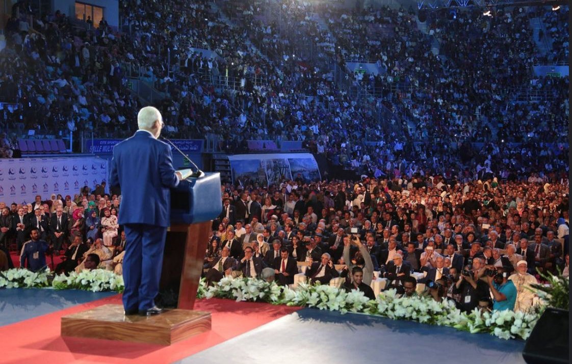 L'image montre un homme debout sur une scène, s'adressant à une foule nombreuse. Il est en costume et se tient derrière un pupitre, probablement en train de donner un discours. La salle est pleine, avec des centaines de personnes assises en face de lui, certaines regardant attentivement. L'éclairage crée une ambiance dramatique, mettant en valeur à la fois le conférencier et le public. Des décorations florales sont visibles au premier plan, ajoutant une touche d'élégance à l'événement.
