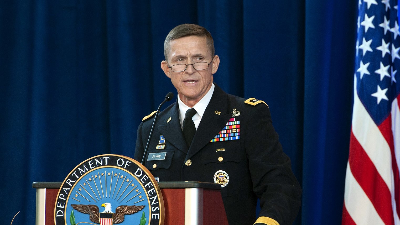 L'image montre un homme en uniforme militaire se tenant derrière un pupitre portant le sceau du département de la Défense des États-Unis. Il semble s'adresser à un public, probablement lors d'une cérémonie officielle ou d'une conférence. Le fond inclut un drapeau américain, ce qui souligne le contexte militaire et gouvernemental de l'image. L'homme a une expression sérieuse et semble engagé dans son discours.