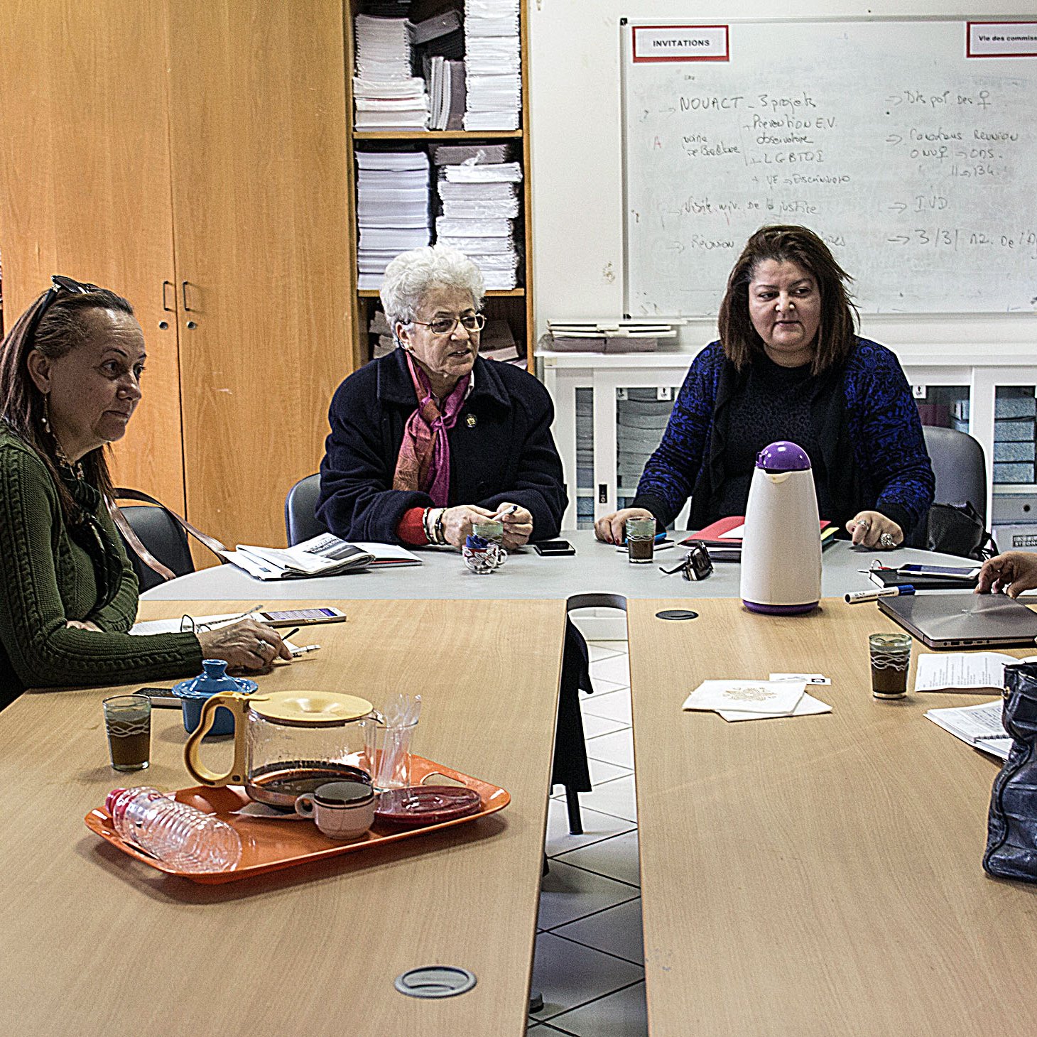 The image depicts a group of five women sitting around a table in a room filled with shelves and papers. They appear to be engaged in a discussion or work-related activity. The table has some items on it, including drinks and papers, suggesting a collaborative atmosphere. The women are looking at each other, indicating communication and interaction among them. The setting seems to be an office or a meeting room.