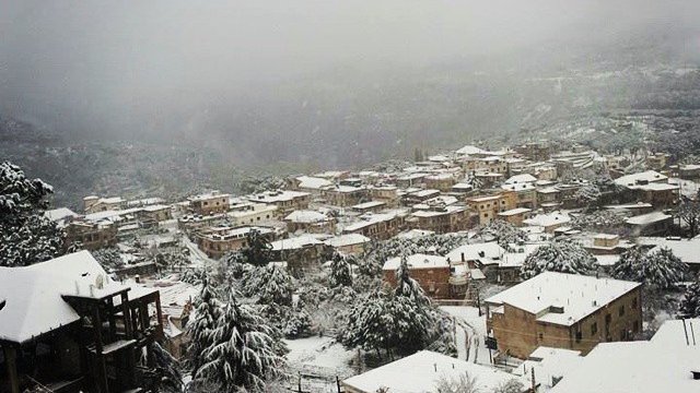 L'image montre un paysage hivernal où une petite ville est recouverte de neige. Les toits des maisons, en grande partie en bois, sont également blancs, créant une atmosphère paisible et froide. On peut apercevoir des arbres chargés de neige et des collines en arrière-plan, le tout enveloppé par une brume légère, ajoutant à la sérénité de la scène. La vue donne une impression de calme et d'isolement, typique des régions montagneuses en hiver.
