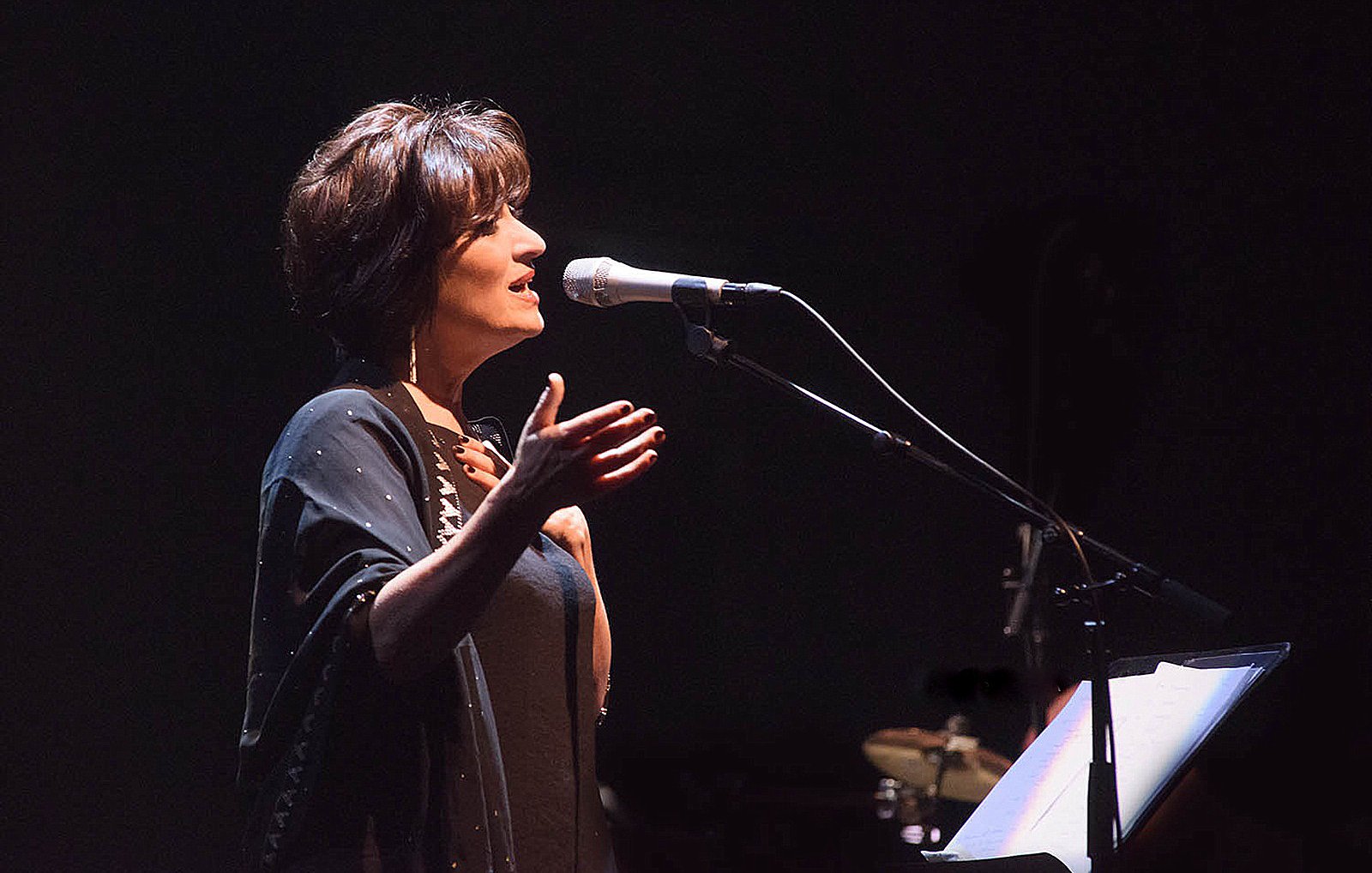 L'image montre une femme sur scène, en train de chanter ou de parler dans un microphone. Elle a les cheveux courts et bruns, et porte une tenue sombre, accompagnée d'un châle. Son expression semble engagée et passionnée, suggérant qu'elle interprète une performance émotionnelle. En arrière-plan, on aperçoit des instruments de musique, ce qui indique qu'elle se produit dans un cadre musical. L'éclairage est tamisé, créant une ambiance intime et captivante.