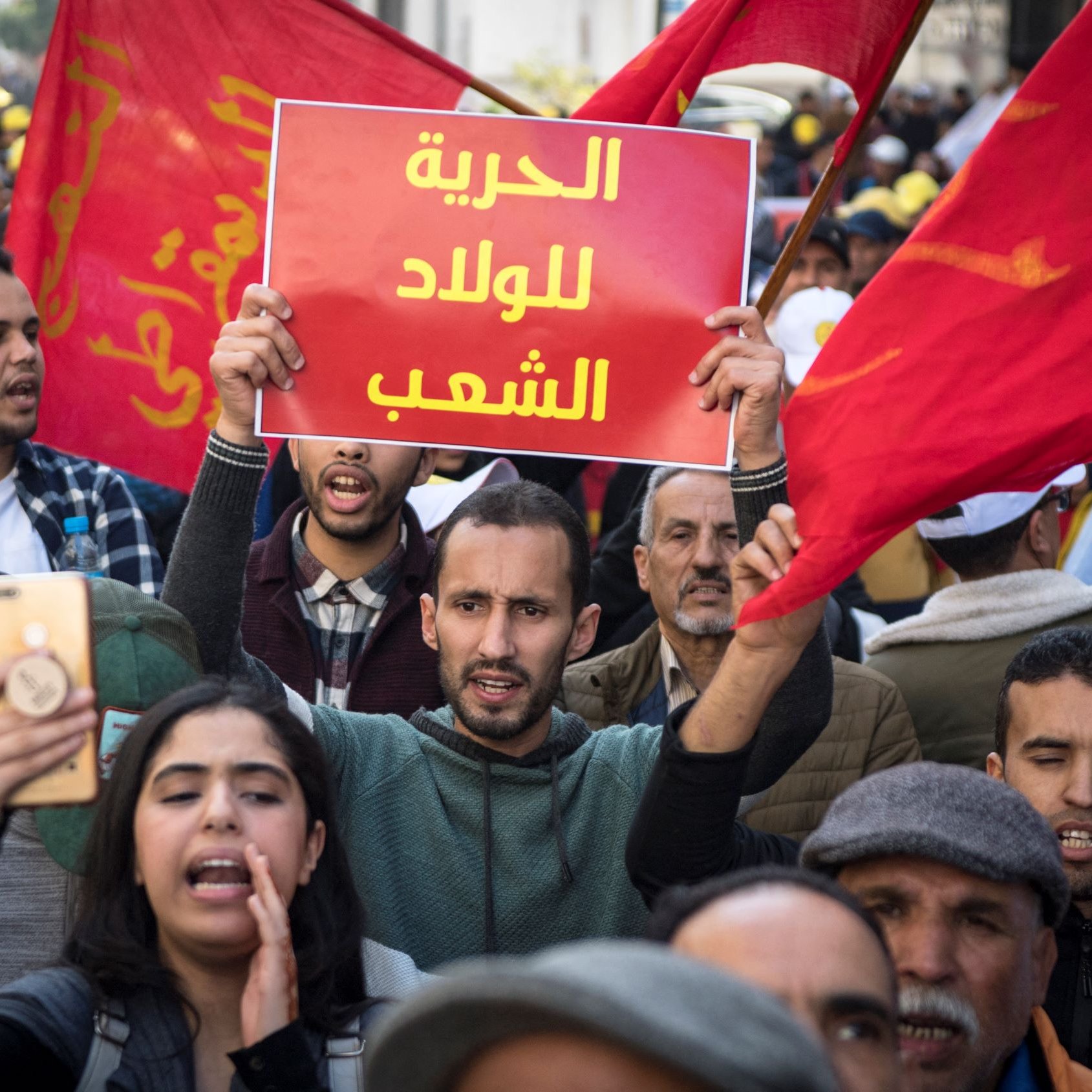 Nell'immagine si vede una manifestazione di protesta affollata, con persone che sventolano bandiere rosse. In primo piano, un giovane tiene in alto un cartello con scritto in arabo "libertà per il popolo". Altre persone intorno a lui sembrano partecipare attivamente, alcune con telefoni in mano per immortalare il momento. L'atmosfera è carica di emozione e determinazione, riflettendo il desiderio di cambiamento e di diritti.