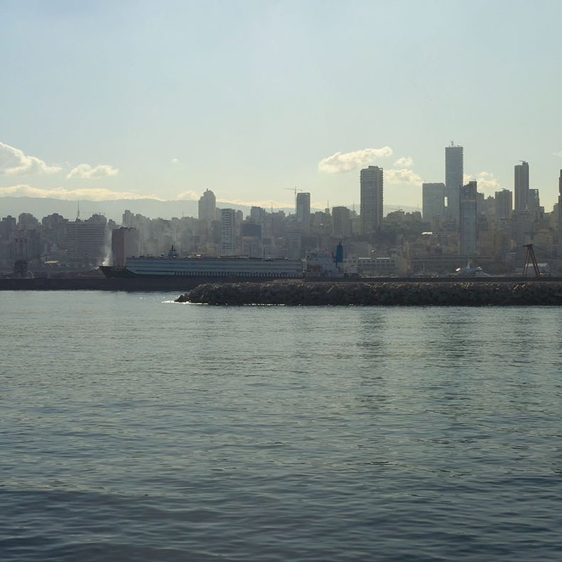 L'image montre un paysage urbain d'une ville moderne en bord de mer. On peut voir des gratte-ciels s'élevant le long de la côte, reflétant la lumière du soleil sur l'eau calme de la mer. Au premier plan, il y a un port ou une jetée, et l'arrière-plan est rempli de bâtiments qui se dressent majestueusement. Le ciel est légèrement nuageux, ce qui crée une ambiance douce et mélancolique. L'ensemble donne une impression de tranquillité et de modernité.
