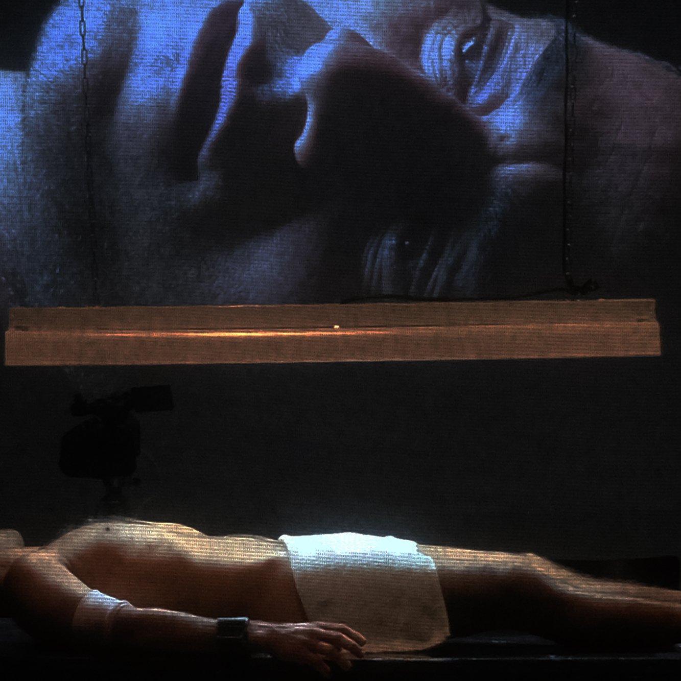 L'image montre un homme allongé sur une surface, partiellement nu, avec une serviette couvrant son corps. Au-dessus de lui, une projection de son visage est affichée sur un grand écran, créant un effet visuel puissant. La lumière provient d'une source suspendue, ajoutant une atmosphère dramatique à la scène. Les détails du visage projeté sont clairement visibles, tandis que l'homme au sol semble dans un état de vulnérabilité. L'ensemble évoque des thèmes de réflexion, d'introspection ou de méditation.