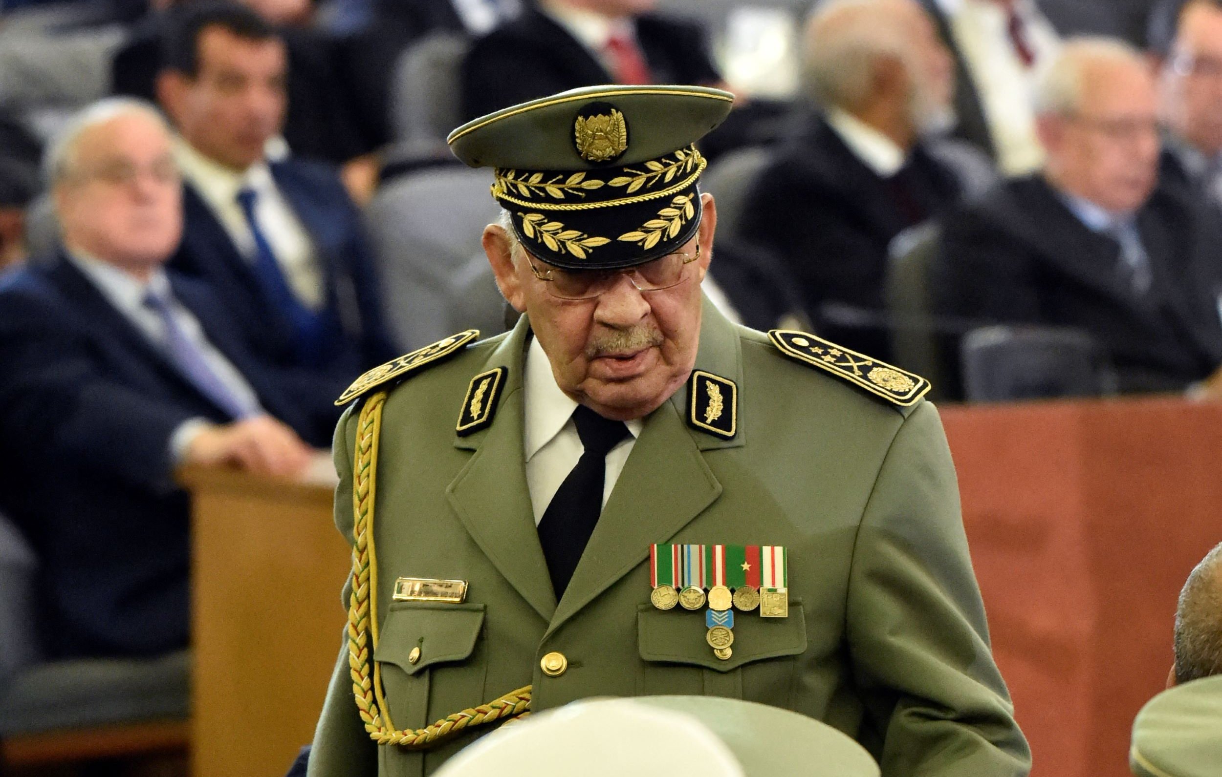 L'image montre un groupe de personnes réunies dans une grande salle, avec des sièges disposés en rangées. Au premier plan, un homme en uniforme militaire avec un chapeau prononcé, orné de médailles, se déplace parmi la foule. Dans l'arrière-plan, on aperçoit d'autres hommes et femmes, certains en costume, d'autres portant des turbans, tous attentifs à l'événement. L'atmosphère semble formelle, suggérant une occasion importante ou une cérémonie.