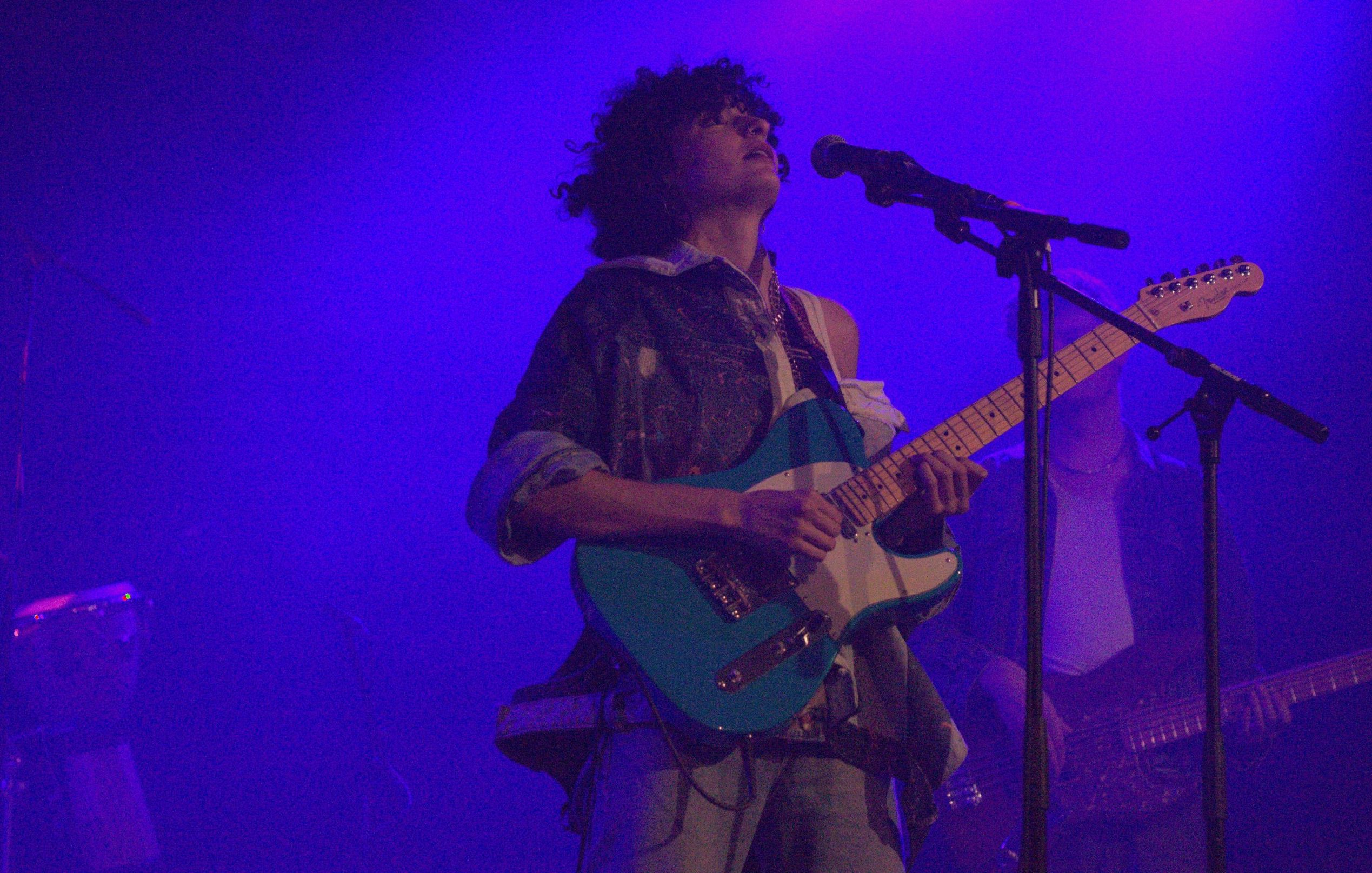 الصورة تظهر عازف جيتار على المسرح. الشخص يرتدي ملابس غير تقليدية ويعزف على آلة الجيتار الكهربائية ذات اللون الأزرق، بينما يبدو أنه يركز في الأداء. الخلفية مضيئة بلون أرجواني، مما يضيف جوًا مميزًا إلى الصورة. يمكن رؤية بعض الآلات الموسيقية الأخرى في الخلفية، مما يشير إلى وجود فرقة موسيقية.