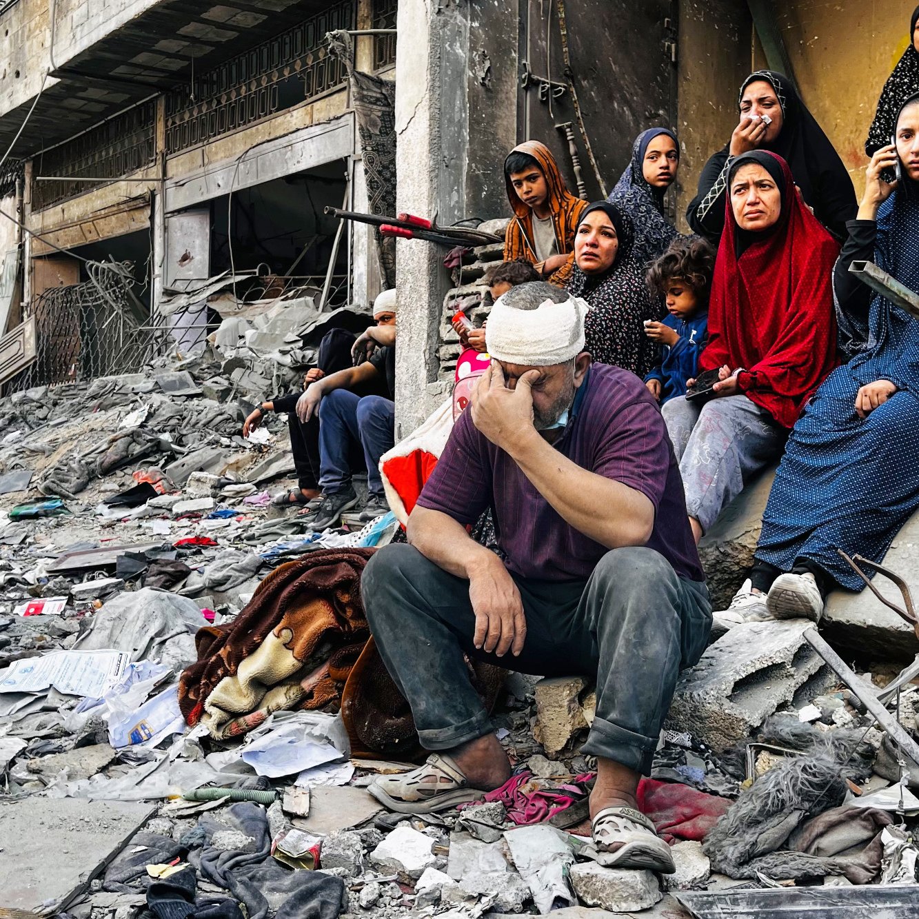 L'image montre un groupe de personnes assises sur des décombres dans une zone de conflit. Au premier plan, un homme, la tête bandée, exprime une grande tristesse ou du désespoir, tandis qu'à proximité, plusieurs femmes en vêtements sombres regardent vers l'objectif, certaines avec une expression préoccupée. Un petit garçon est assis un peu plus loin, semblant perdu dans ses pensées. L'environnement est chaotique, avec des ruines et des débris autour, suggérant des dommages importants causés par des événements récents. L'atmosphère générale est lourde, reflétant la douleur et la résilience des personnes présentes.