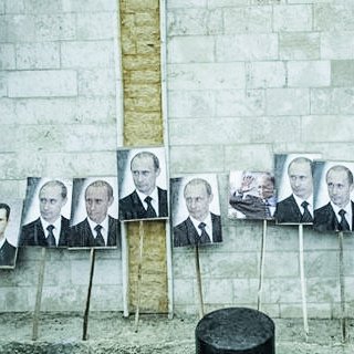 L'image montre un mur en béton avec plusieurs portraits accrochés, principalement des visages d'un homme en costume. Ces portraits sont placés sur des supports en bois. Une personne, dont on ne voit que le dos, marche à proximité. Le décor semble urbain et pourrait impliquer un contexte politique. Les expressions faciales sur les portraits sont sérieuses, reflétant une ambiance de propagande ou de soutien.