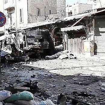 L'image montre une rue dévastée, probablement dans une zone de conflit. On peut voir des bâtiments endommagés et détruits, avec des débris éparpillés sur le sol. Des sacs de sable sont accumulés sur le côté, ce qui suggère des efforts de protection. Un panneau de signalisation est visible, mais l'environnement semble plutôt déserté et en ruines. L'atmosphère dégage un sentiment de désolation et de destruction.