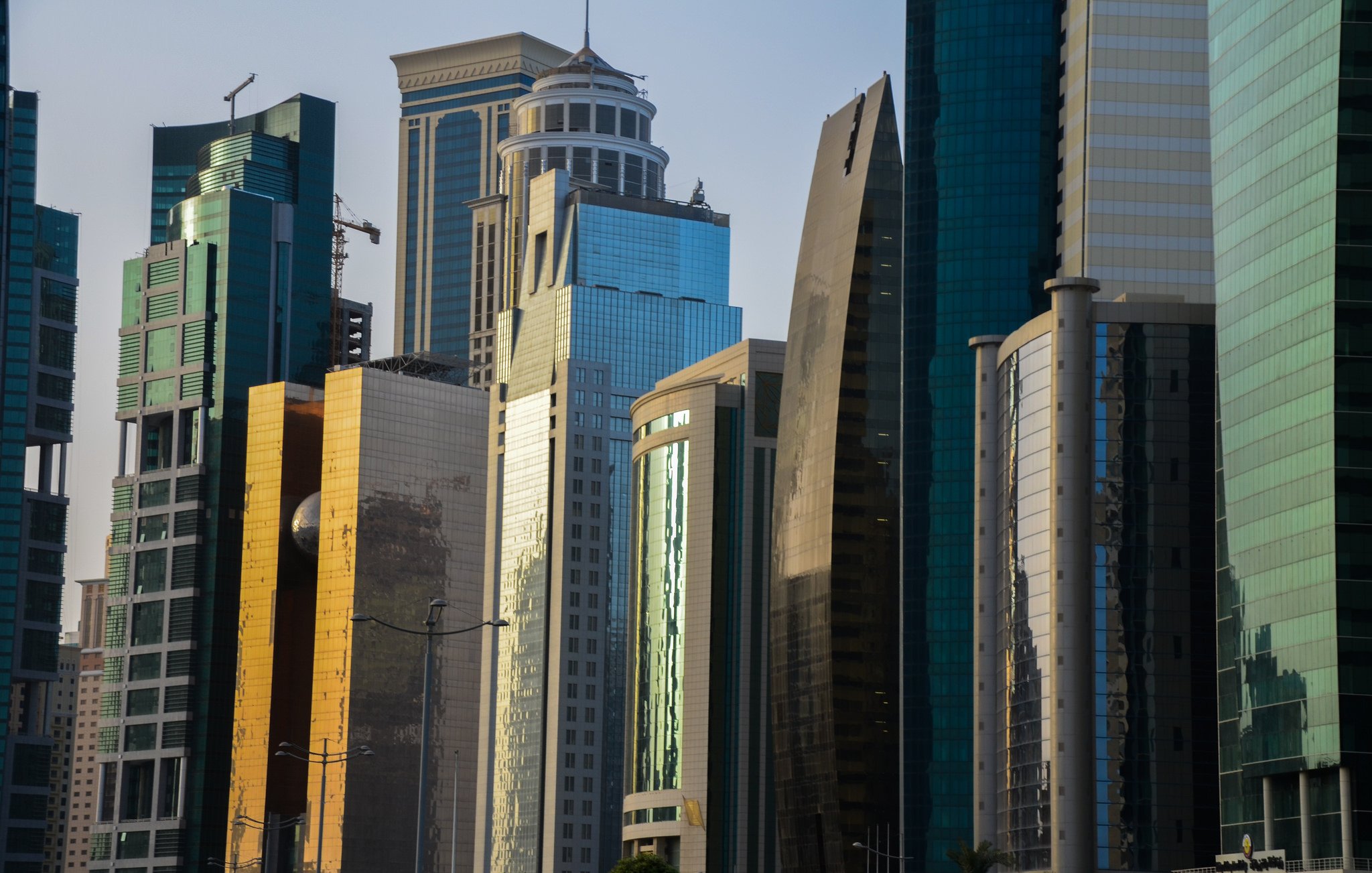 الصورة تظهر مجموعة من ناطحات السحاب الحديثة في منطقة حضرية. تتميز المباني بتصاميمها المعمارية المتنوعة، حيث تختلف في الارتفاع والأشكال. الواجهة الزجاجية لبعض المباني تعكس الضوء مما يعطيها لمسة عصرية. الألوان تتراوح بين الزجاج الأزرق والأخضر، مع وجود لمسات من الذهبي والرمادي. السماء في الخلفية توحي بأوقات النهار، مما يضفي جواً حيوياً على المشهد.