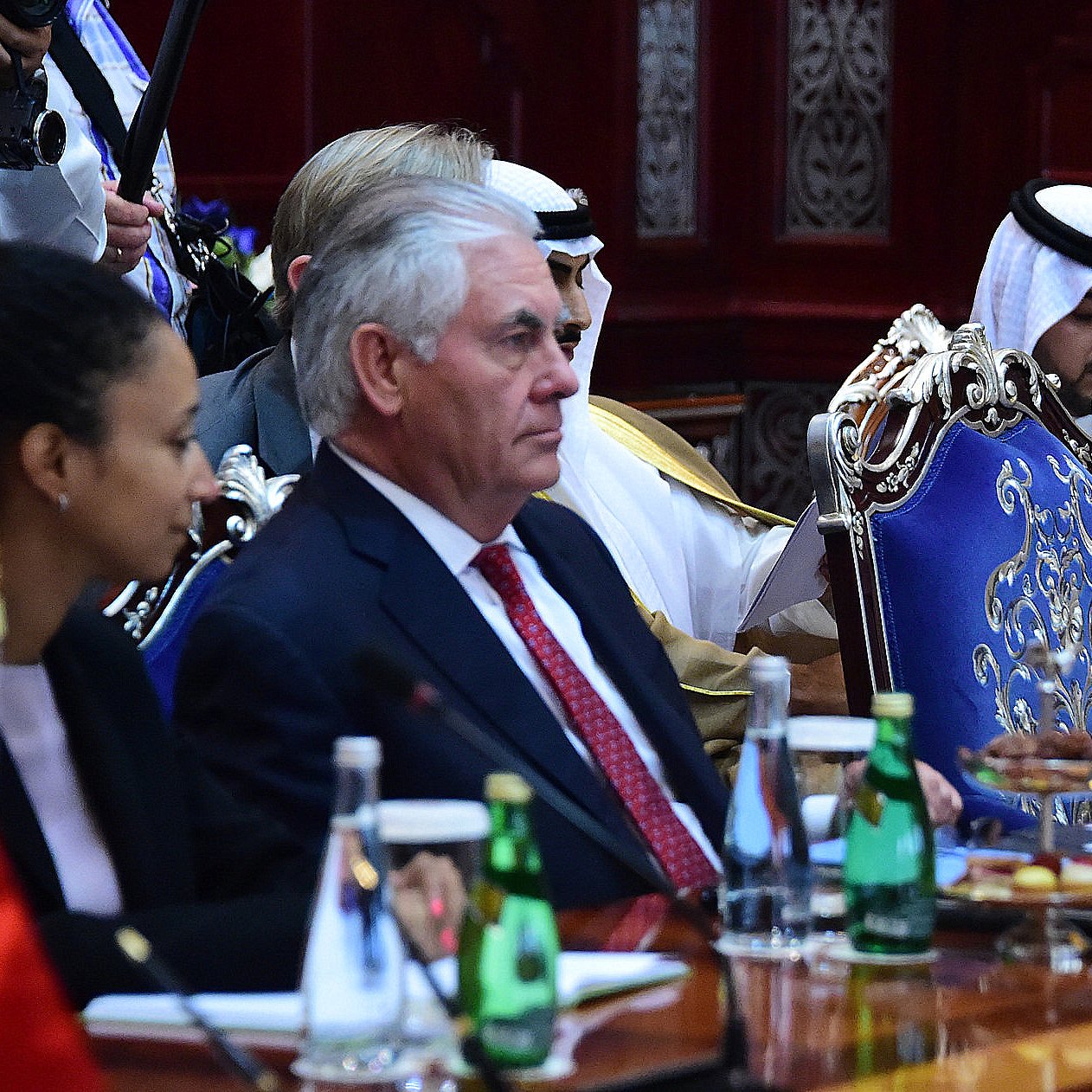L'image présente une réunion officielle, où l'on peut voir plusieurs personnes assises autour d'une grande table. À gauche, une femme est accoudée à la table, tandis qu'à droite, un homme en costume, probablement un diplomate, semble écouter attentivement. Au centre, il y a un drapeau américain, et derrière, d'autres participants portant des vêtements traditionnels peuvent être aperçus. L'environnement semble formel, avec une décoration riche et des bouteilles d'eau sur la table.