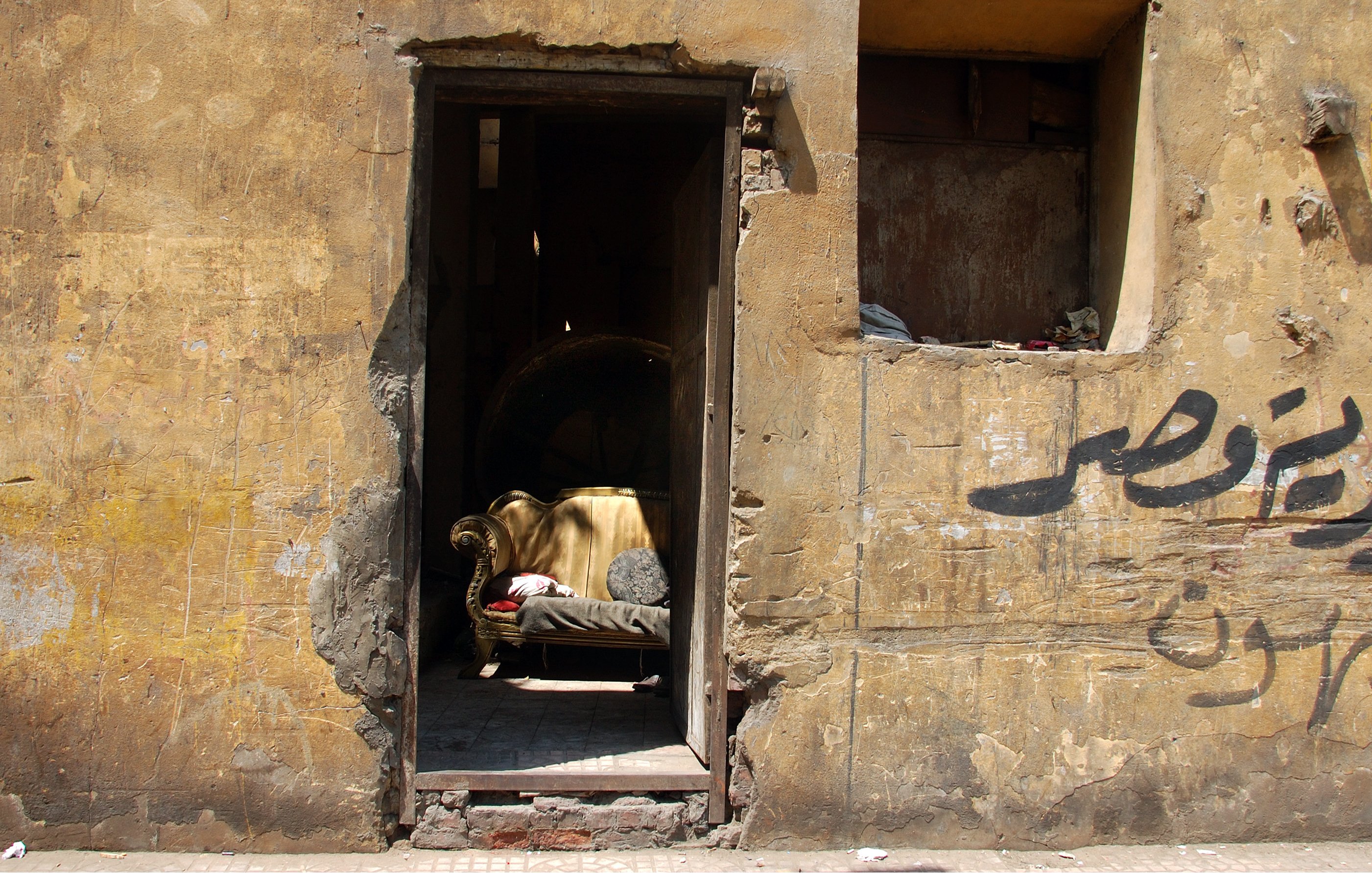 L'image montre une entrée partiellement ouverte d'un bâtiment avec un mur aux teintes jaunies et en mauvais état. À l'intérieur, on distingue un canapé doré, qui semble être un élément de mobilier abandonné. Le mur présente aussi des inscriptions en arabe, ajoutant une touche culturelle à la scène. L'ensemble dégage une atmosphère nostalgique, évoquant un espace autrefois habité et maintenant oublié.