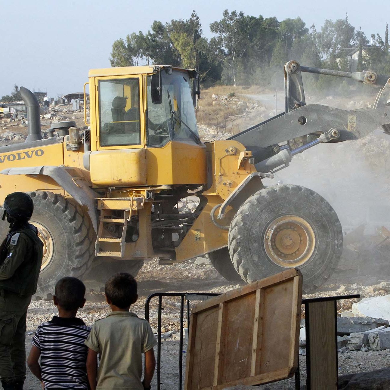 L'image montre un camion-tracteur de type Volvo en action, probablement en train de déblayer ou de détruire des structures. En premier plan, deux jeunes enfants observent la scène. À l'arrière-plan, on peut voir des débris et des bâtiments partiellement détruits, ainsi qu'un soldat en tenue militaire. L'atmosphère semble tendue, avec de la poussière soulevée par le véhicule.