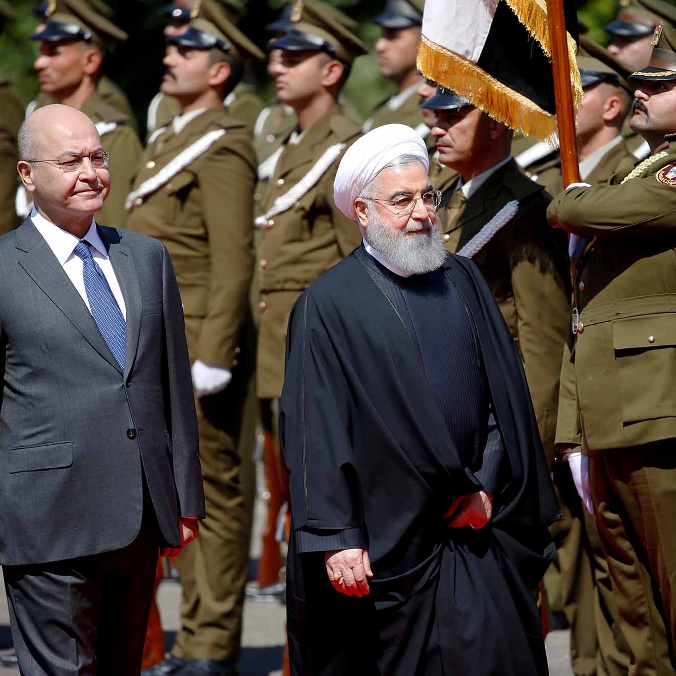 L'image montre deux hommes en tenue officielle, se tenant côte à côte lors d'une cérémonie. L'un est vêtu d'un costume élégant, tandis que l'autre porte une robe traditionnelle sombre avec une coiffure distinctive. En arrière-plan, des militaires en uniforme se tiennent au garde-à-vous, tenant des drapeaux. Le décor semble être une cérémonie officielle ou un événement diplomatique.