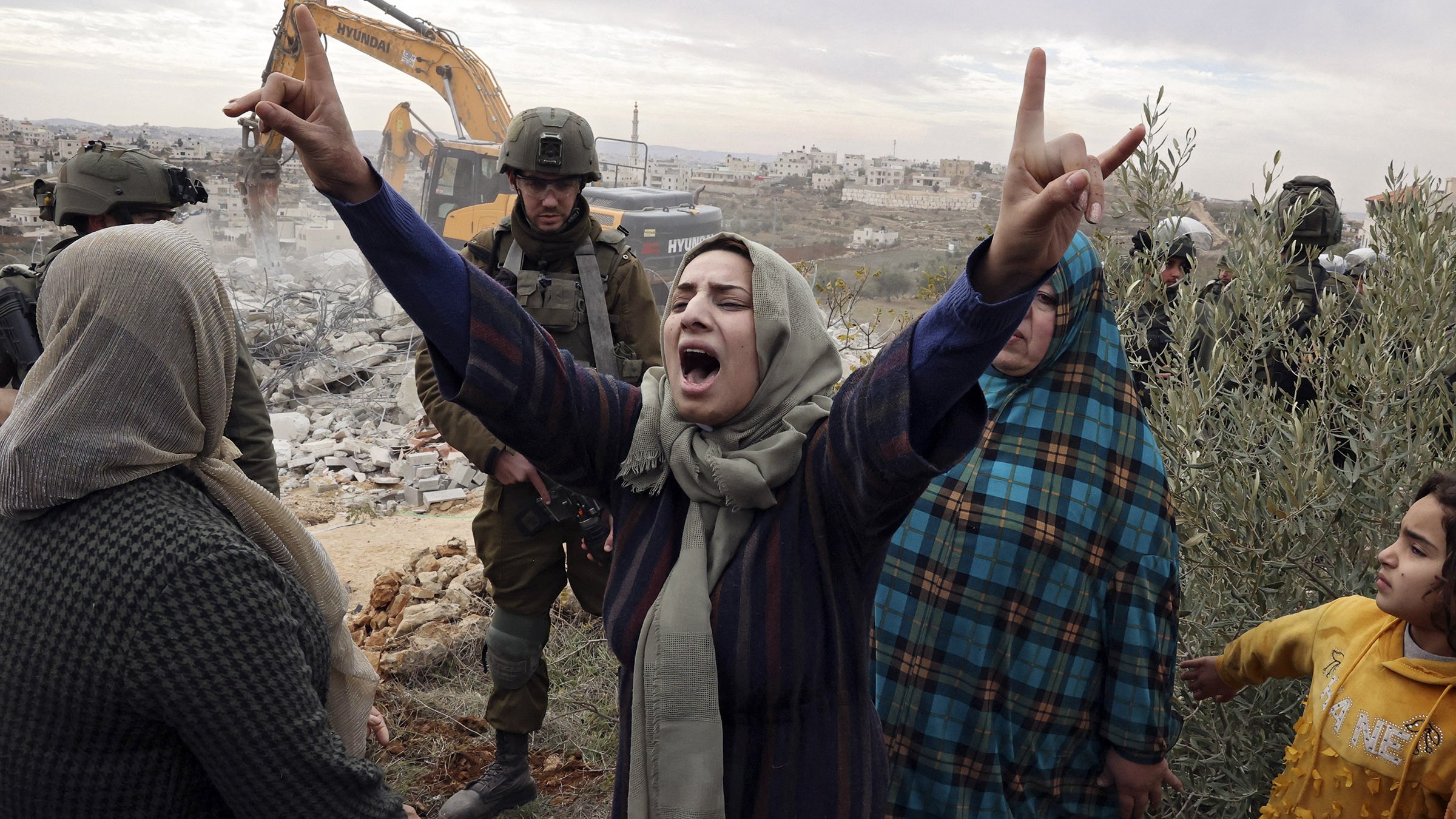 L'image montre une scène intense où une femme exprime sa colère et sa frustration, levant les bras en signe de protestation. Elle est entourée de plusieurs personnes, dont certaines ont l'air préoccupées ou attristées. En arrière-plan, on aperçoit des forces de l'ordre et un engin de construction qui semble démolir une structure. L'atmosphère dégagée par l'image est chargée d'émotion, suggérant des tensions et des luttes en cours.