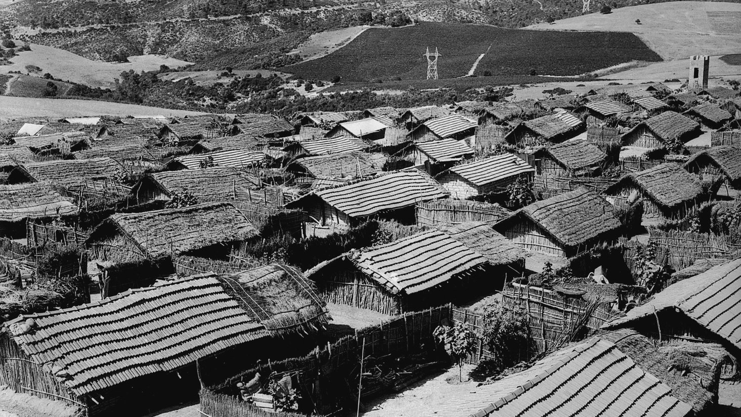 الصورة تُظهر منظرًا عامًّا لقرية صغيرة تتكون من مجموعة من المنازل التقليدية. المنازل تبدو مصنوعة من مواد طبيعية مثل الخشب والقش، مع أسطح مائلة. يتواجد حول القرية تلال خضراء ومناظر طبيعية جميلة، مما يضفي على المشهد طابعًا ريفيًا هادئًا. السماء تبدو صافية، مما يبرز جمال المكان.