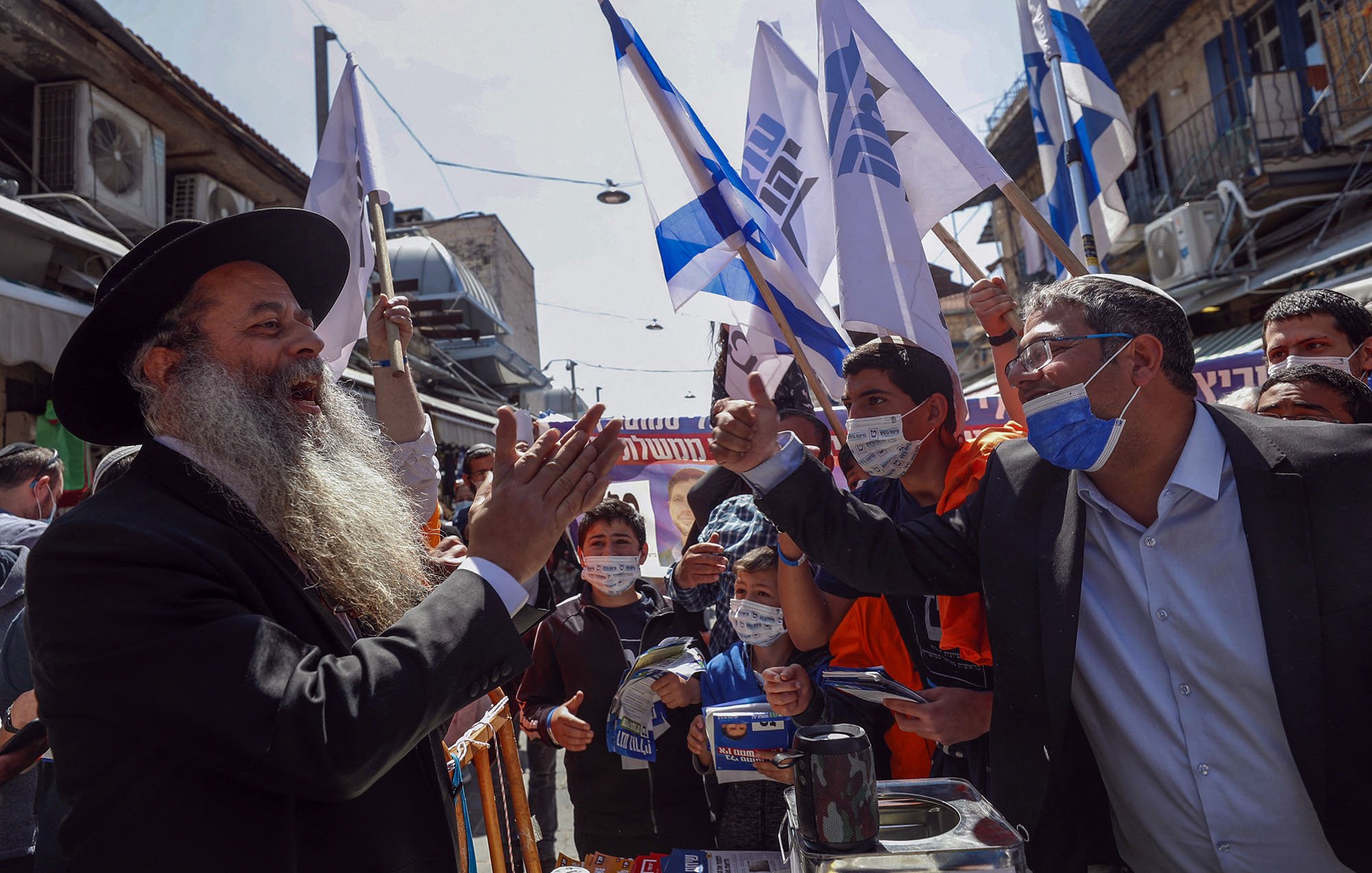 L'image montre une scène animée dans une rue, probablement lors d'un rassemblement ou d'une manifestation. Des personnes se tiennent debout, brandissant des drapeaux, apparemment en soutien à une cause. Au centre, un homme avec une longue barbe et un chapeau noir s'adresse à un autre homme, qui semble engagé dans un échange passionné. Les deux semblent entourés par une foule de jeunes gens, certains portant des masques de protection. L'environnement urbain et les bâtiments en arrière-plan ajoutent à l'atmosphère dynamique de l'événement.