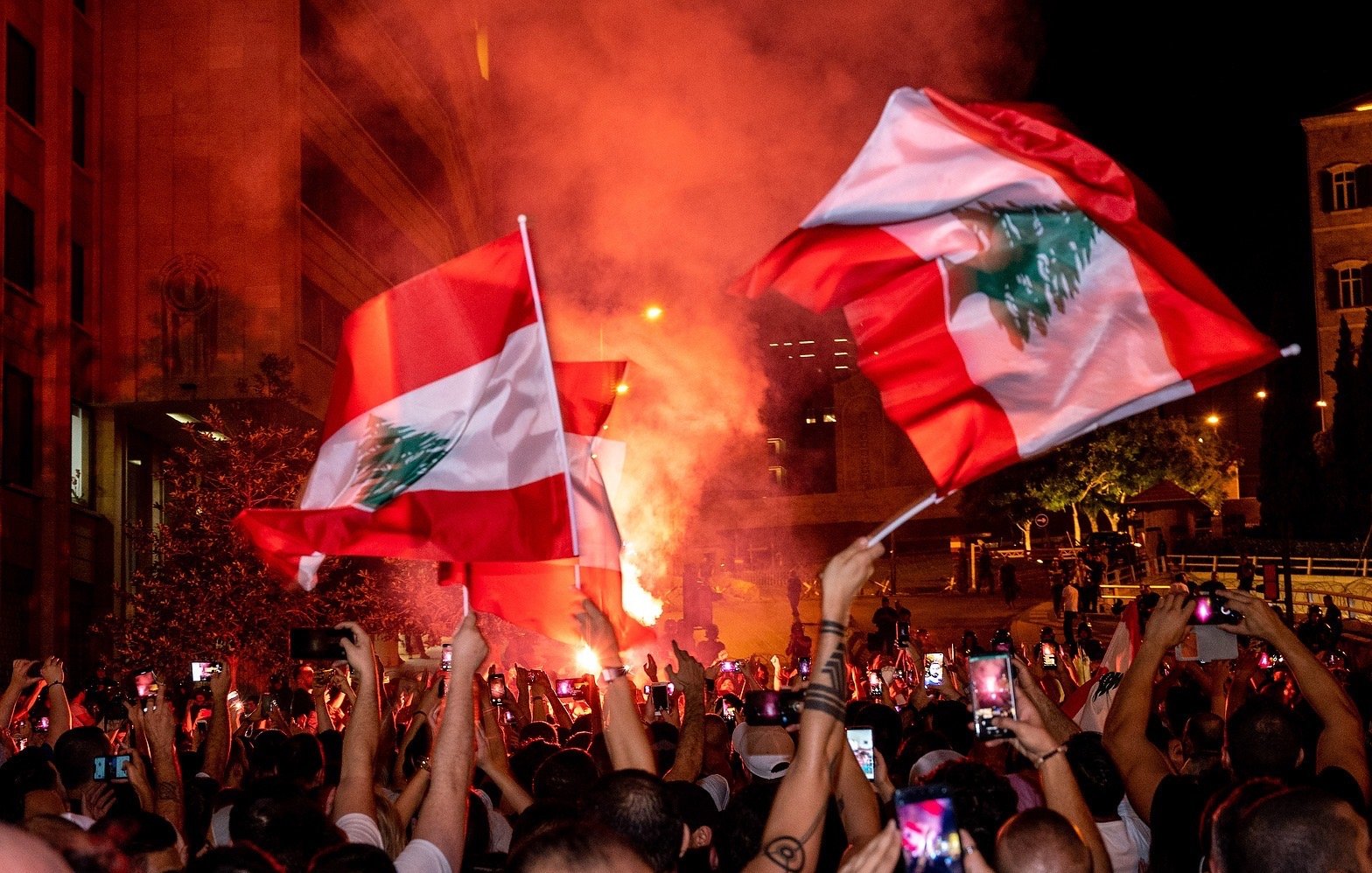 در تصویر، جمعیتی از مردم در یک تظاهرات شبانه دیده می‌شوند. افراد با پرچم‌های لبنان در دست، در حال شادی و ابراز احساسات هستند. یک شعله آتش بزرگ در پس‌زمینه به روشنایی فضای شب افزوده و به جو فعالیت و هیجان دامن می‌زند. تعداد زیادی از افراد گوشی‌های هوشمند خود را برای ثبت لحظات بالا برده‌اند. این صحنه نمایانگر شور و شوق اجتماعی و همبستگی مردم است.