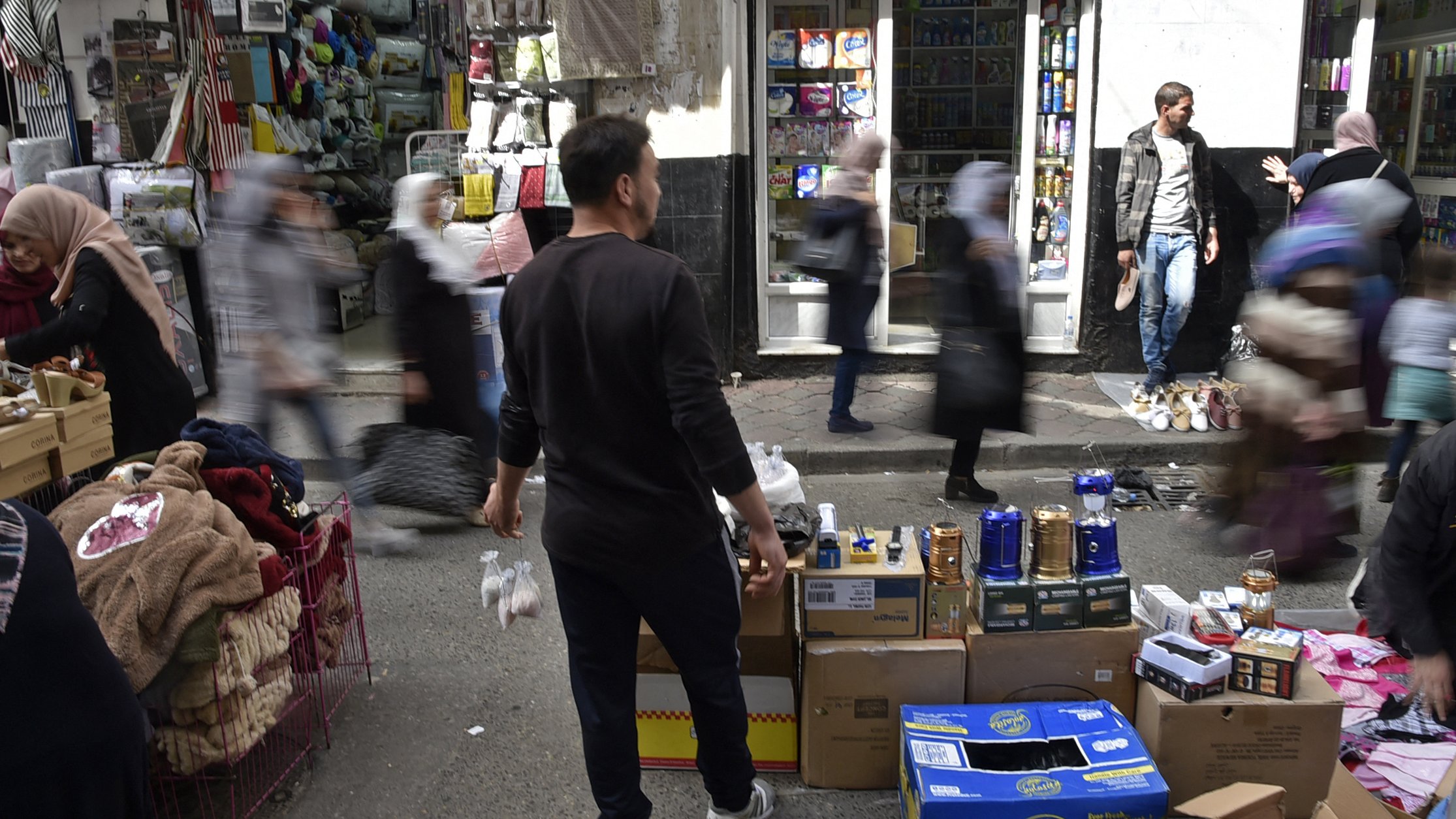 L'image montre une scène animée d'un marché urbain. On peut voir plusieurs personnes se déplacer rapidement, créant un effet de flou. Au premier plan, un homme se tient de dos, tenant des objets. Des étals sont visibles, avec des boîtes et des marchandises disposées sur des chariots. En arrière-plan, des magasins sont ouverts et on aperçoit des clients. L'atmosphère semble vivante et dynamique, typique d'un marché fréquenté.