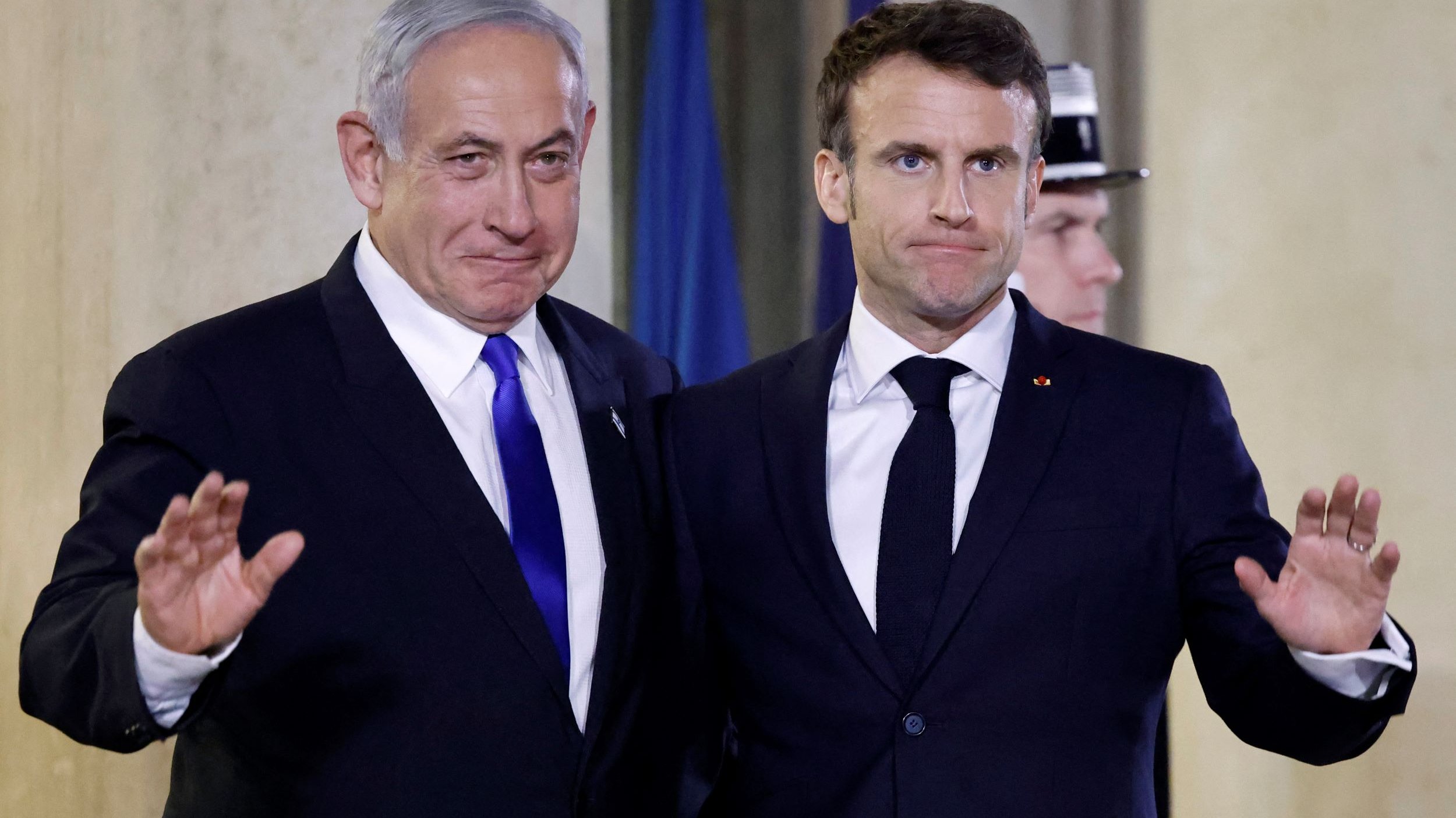 Nell'immagine si vedono due uomini in abiti formali, sorridenti e in posa per una foto. Sono entrambi in piedi, con una mano sollevata in segno di saluto. Lo sfondo presenta un ambiente elegante, probabilmente all'interno di un edificio istituzionale. Gli uomini sembrano essere in un contesto ufficiale, come una cerimonia o un incontro diplomatico.