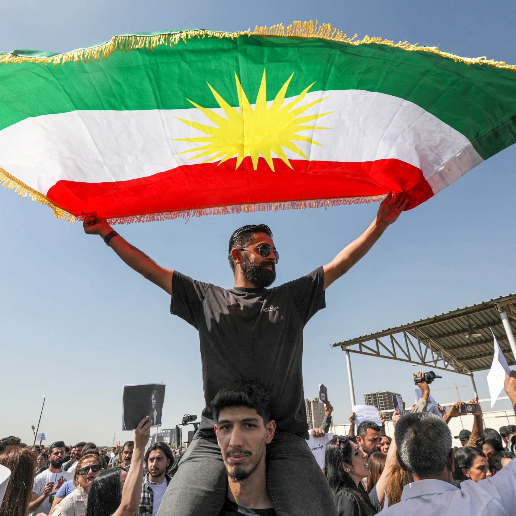 L'image montre un groupe de personnes lors d'une manifestation. Au premier plan, un homme se tient sur les épaules d'un autre, brandissant un grand drapeau kurde, qui est composé de bandes vertes, rouges et blanches avec un soleil jaune au centre. La foule autour d'eux paraît enthousiaste et engagée. Le ciel est clair, ce qui indique une journée ensoleillée. Des pancartes sont également visibles parmi les manifestants, suggérant qu'ils expriment des revendications ou des support pour une cause.