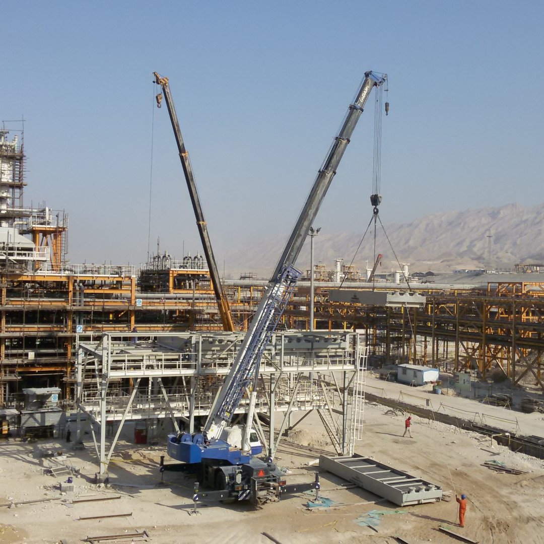 L'image montre un chantier de construction avec plusieurs grues sur le site. On peut voir des structures métalliques en voie d'achèvement à l'arrière-plan, ainsi que des véhicules de chantier et des ouvriers sur le site. Le paysage environnant est montagneux, et le ciel est dégagé, ce qui suggère une journée ensoleillée. Le chantier semble être à un stade de construction avancé, avec des matériaux disposés sur le sol.