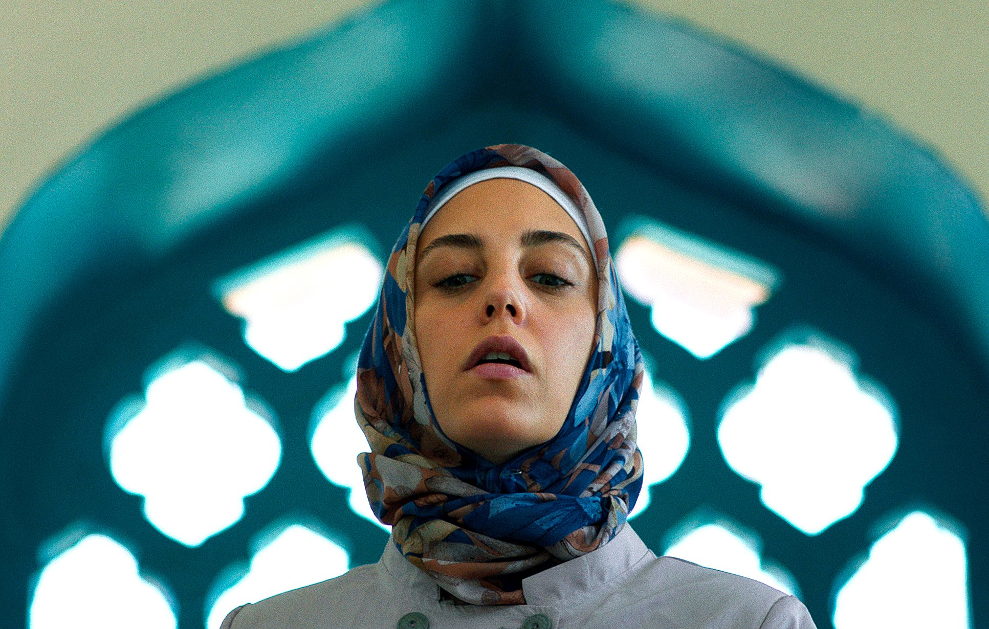 L'image montre une femme portant un hijab coloré, se tenant dans un cadre architectural. La lumière met en valeur son visage et le fond présente des motifs géométriques typiques des designs islamiques, avec des arches et des vitraux. Son expression est sérieuse, ce qui ajoute à la profondeur de la scène.