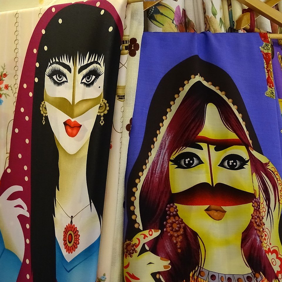 L'image présente deux illustrations de femmes, affichées sur des tissus. À gauche, une femme porte un foulard avec des dessins floraux, des yeux accentués et une bouche rouge. À droite, une autre femme arbore des bijoux traditionnels et un voile coloré, avec un visage stylisé et des traits distinctifs. Les deux illustrations ont un style graphique vibrant et coloré, avec des motifs qui ajoutent à leur allure artistique.