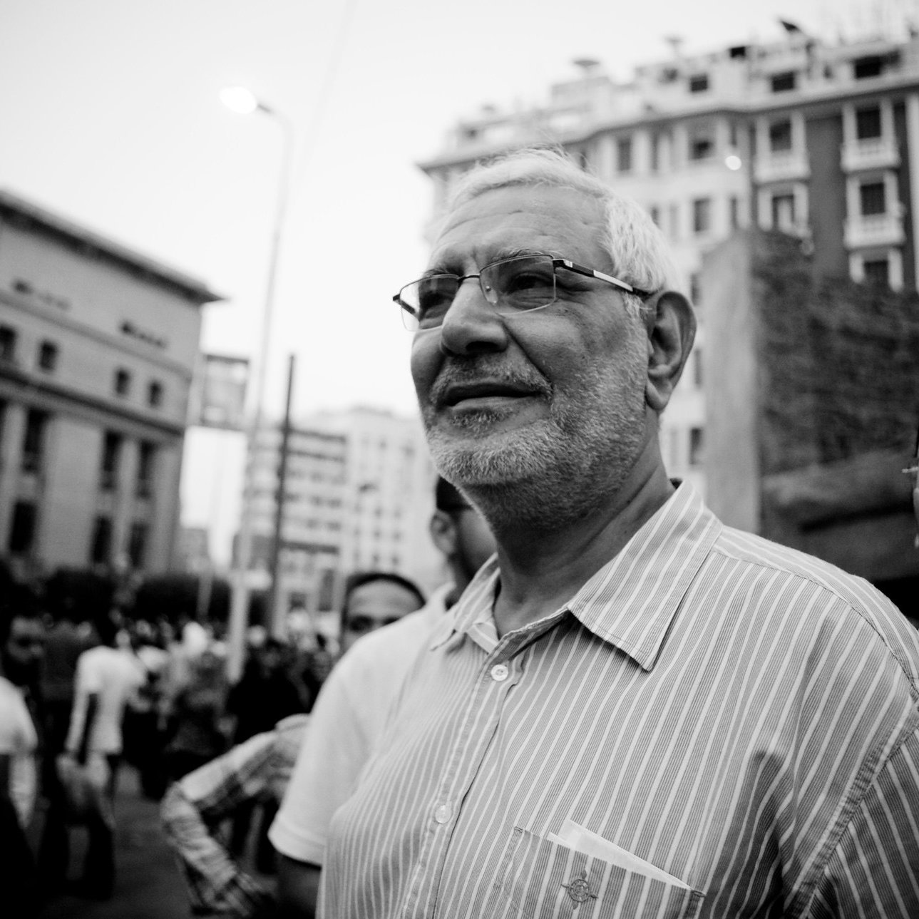 الصورة تظهر رجلًا مسنًا يرتدي نظارات وقميصًا مخططًا، يبدو مبتسمًا ويتحدث مع مجموعة من الأشخاص في خلفيته. الجو العام يوحي بوجود تجمع أو حدث اجتماعي، حيث تُظهر المباني العالية خلفه أجواء حضرية. الصورة بالأبيض والأسود، مما يضيف لمسة درامية للمشهد.
