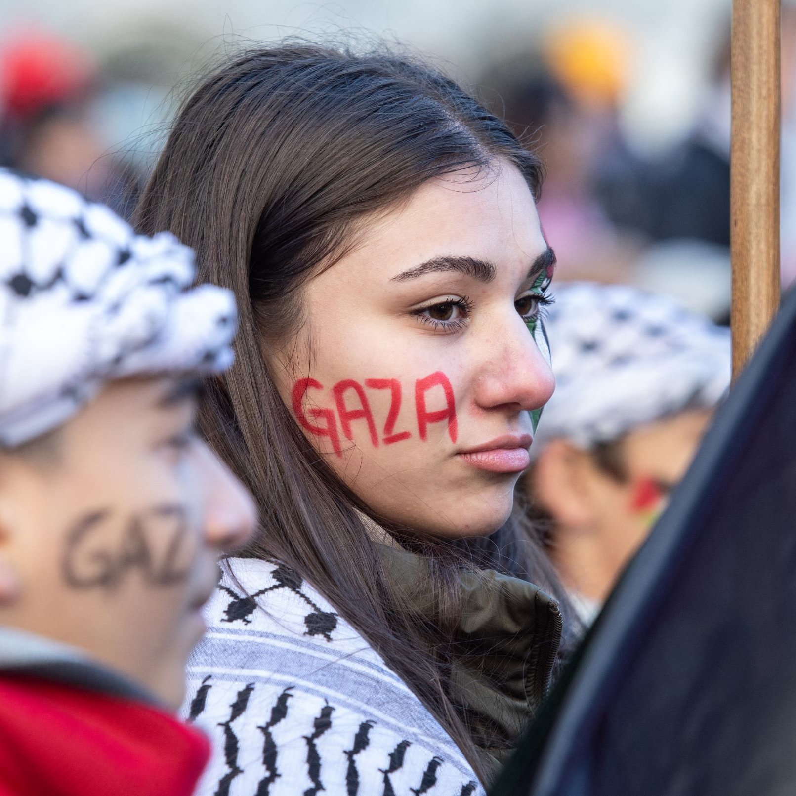 L'image montre des manifestants participant à une manifestation, probablement en lien avec la situation à Gaza. Une jeune femme a le mot "Gaza" peint en rouge sur sa joue, tandis qu'une autre personne à côté d'elle porte un foulard traditionnel palestinien et a également "Gaza" écrit sur son visage, mais en noir. Ils sont entourés d'un public qui semble mobilisé pour une cause. L'atmosphère est sérieuse, reflétant un engagement fort pour la situation en Palestine.