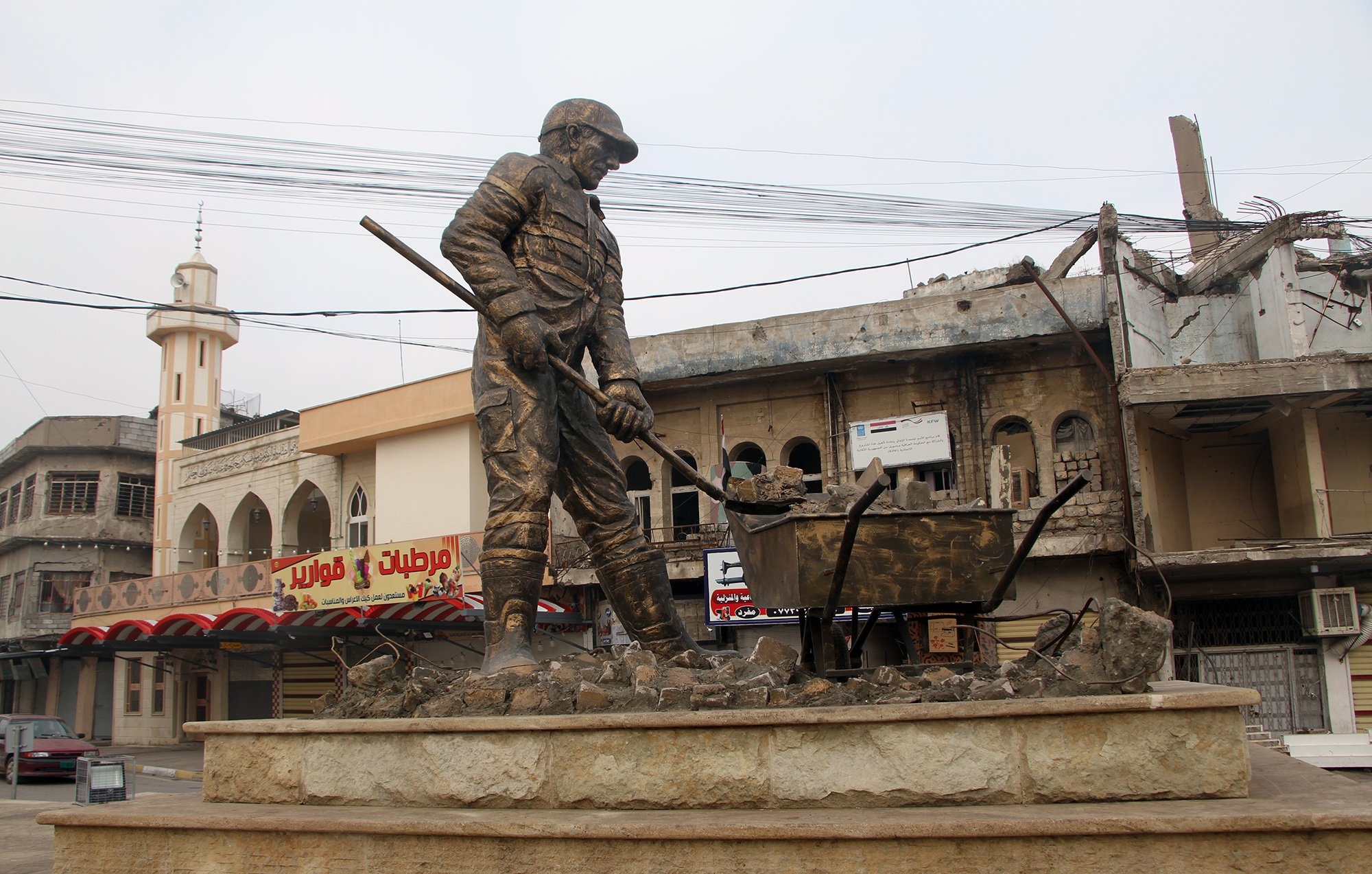L'immagine mostra una statua di un operaio che lavora, con una pala in mano e una carriola accanto a lui. Il monumento è situato in una zona urbana, caratterizzata da edifici danneggiati e in rovina sullo sfondo. La scena trasmette un senso di resilienza e dignità, riflettendo la vita e il lavoro in un contesto difficile. L'atmosfera è un po' desolata a causa delle strutture danneggiate, mentre la statua rappresenta il duro lavoro e l'impegno quotidiano delle persone.