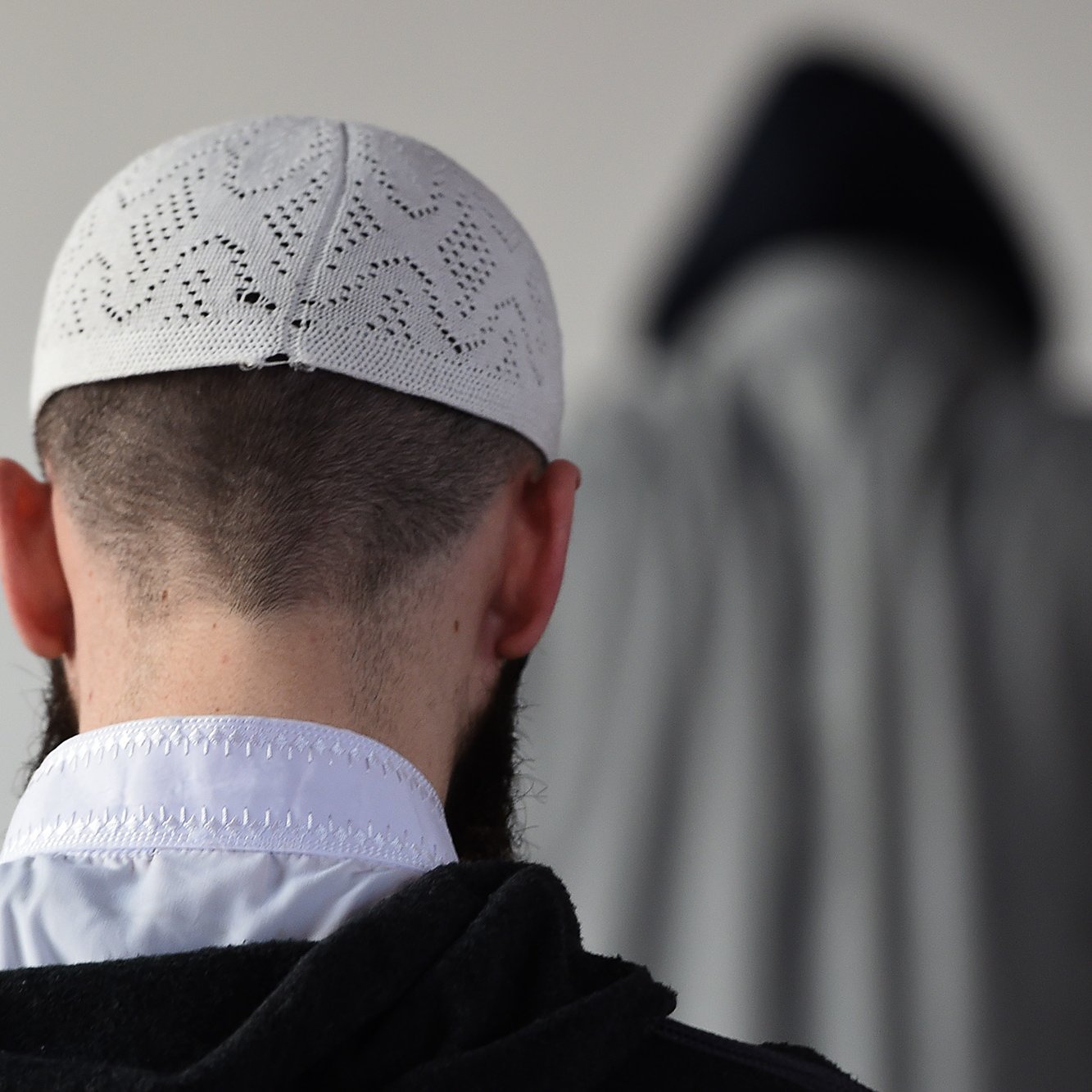 L'image montre un homme de dos, portant un taqiyah (un couvre-chef traditionnel), avec une barbe visible. En arrière-plan, on peut apercevoir une silhouette floue d'une autre personne, vêtue d'une cape ou d'un vêtement similaire. L'environnement semble simple et neutre, avec peu d'éléments distrayants. La scène évoque une ambiance calme et contemplative.