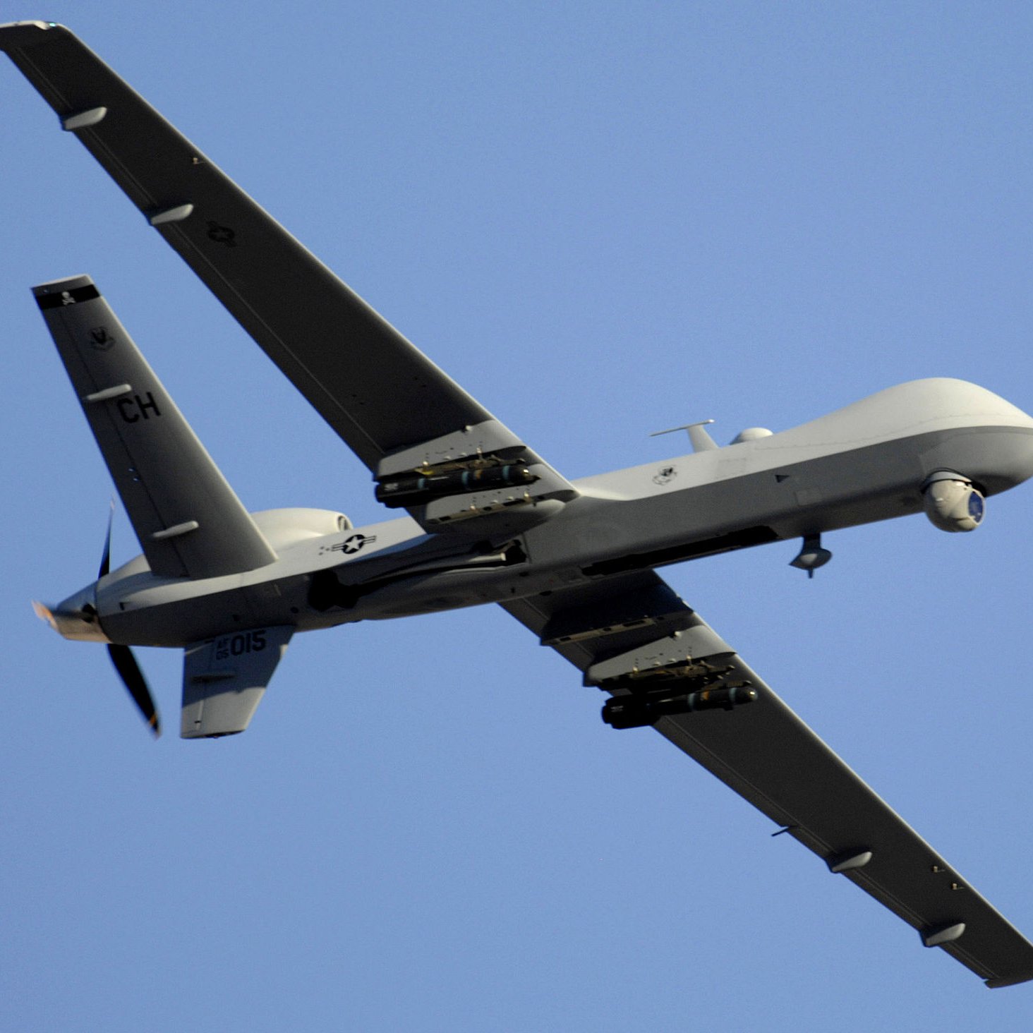 L'image montre un drone de type militaire, en vol. Il a une conception aérodynamique avec des ailes étendues et une queue distinctive. La surface du drone est peinte en gris, ce qui le rend moins visible en altitude. On peut également observer des équipements sous ses ailes, probablement destinés à la surveillance ou à des missions de reconnaissance. Le ciel en arrière-plan est dégagé, mettant en valeur le drone en mouvement.