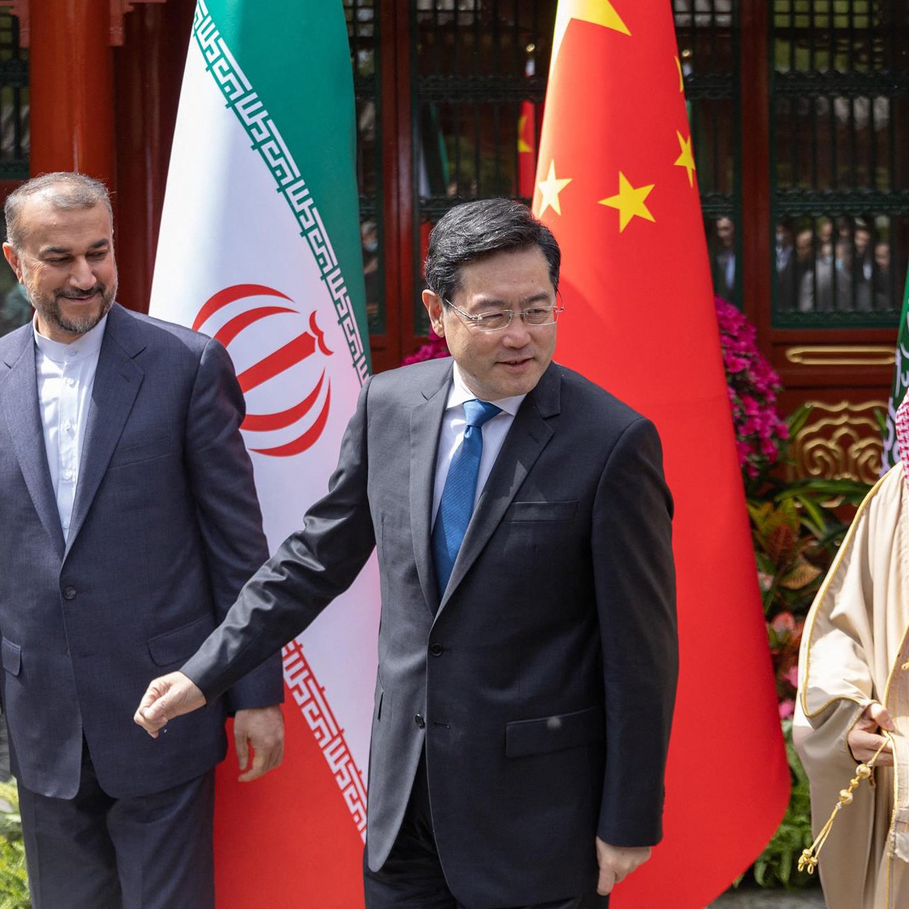 L'immagine mostra tre uomini che si trovano in un contesto formale e diplomatico, con bandiere dell'Iran, della Cina e dell'Arabia Saudita in background. I soggetti sembrano essere coinvolti in una discussione o in un incontro. L'atmosfera sembra prestigiosa e rappresenta un momento di incontro tra le nazioni.
