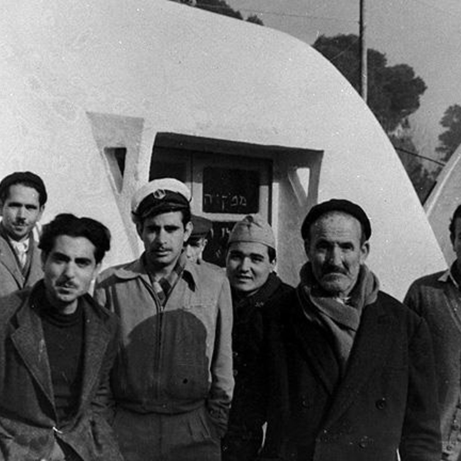 L'image montre un groupe de six hommes, debout près d'une structure blanche ressemblant à une maison ou une cabane. Ils sont habillés de manière variée, certains portant des chapeaux et des manteaux, ce qui suggère un temps frais. L'arrière-plan présente une autre structure similaire et des arbres flous. L'atmosphère semble décontractée, mais les expressions des hommes sont sérieuses.