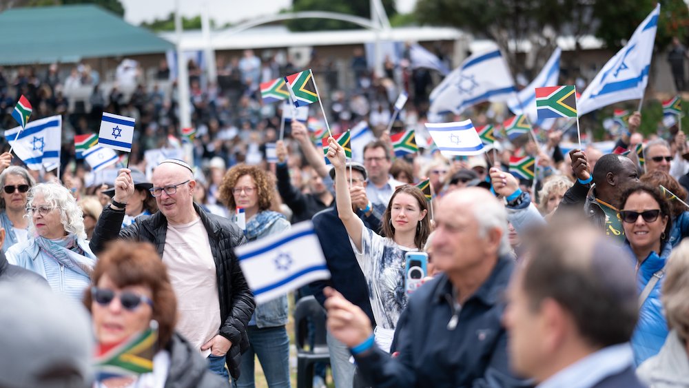 L'image montre une grande foule rassemblée, tenant des drapeaux israéliens et sud-africains. Les personnes semblent engagées et heureux, certaines souriant et levant les drapeaux en signe de soutien. L'environnement est un espace extérieur, et on peut voir des stands ou des tribunes en arrière-plan, suggérant un événement public ou une cérémonie. L'atmosphère semble festive et solidaire.
