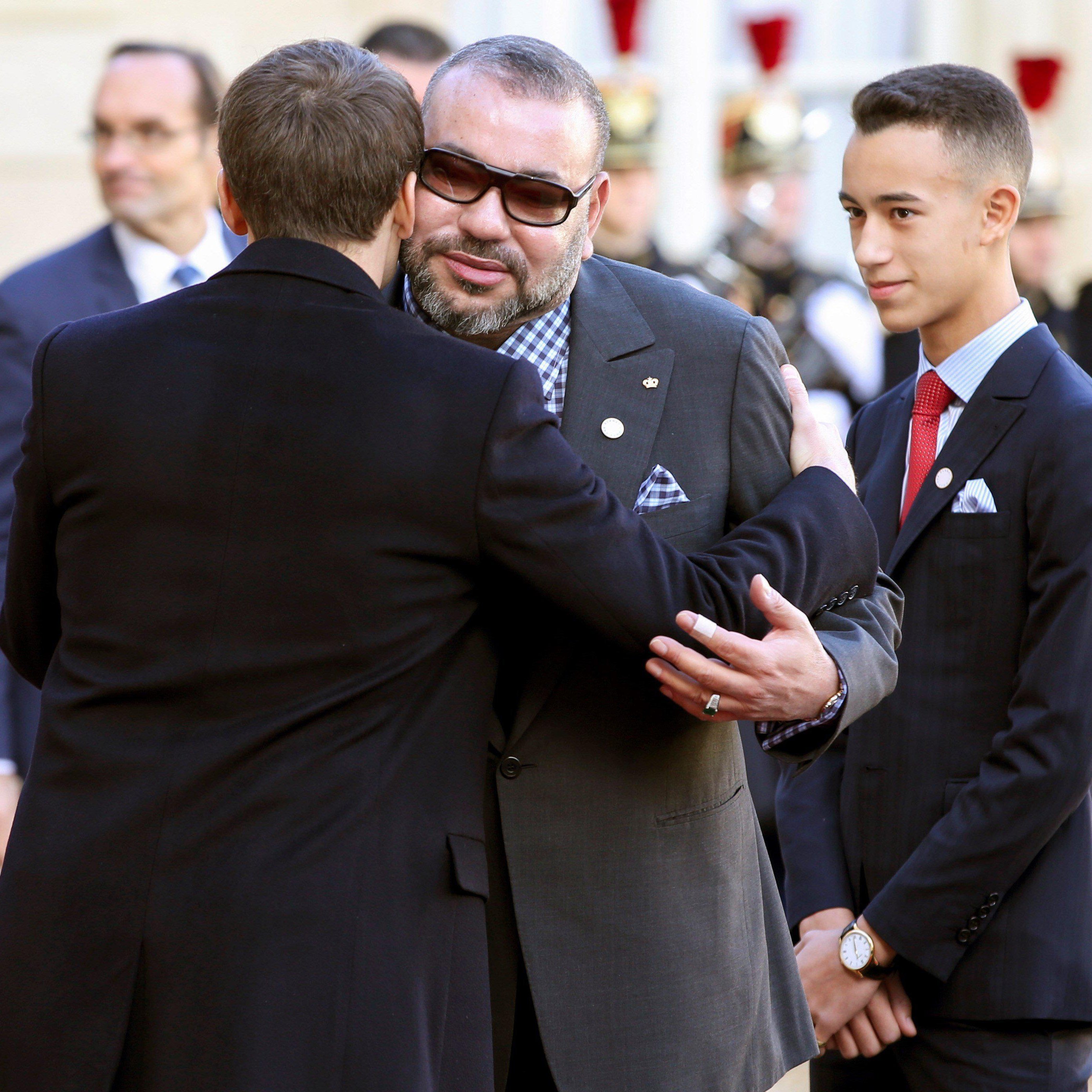 تظهر الصورة مشهدًا رسميًا يتضمن شخصية بارزة تحتضن شخصية أخرى، بينما يقف شاب بجانبهم. الشخص الذي يتم احتضانه يبدو وكأنه يرتدي نظارات شمسية وبدلة أنيقة، ويبدو أنه يتلقى التحية بطريقة ودية. خلفهم، يوجد عدد من أفراد الحرس يرتدون زيًا رسميًا، مما يعكس الطابع الرسمي والمناسبات الدبلوماسية. الجو العام يعكس الاحترام والتقدير المتبادل بين الشخصيات.